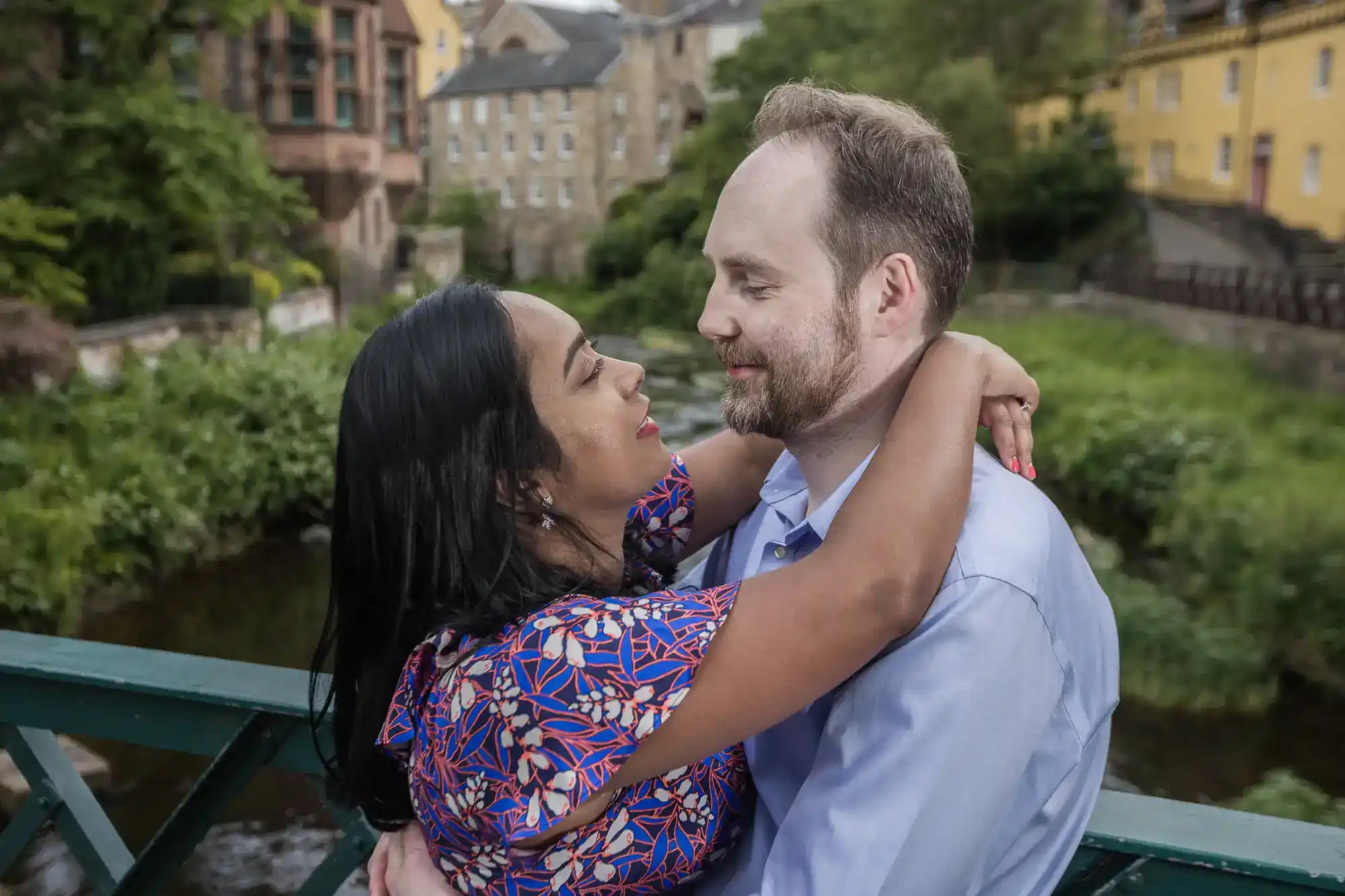 A couple stands on a bridge, embracing and looking into each other's eyes, with greenery and buildings in the background.