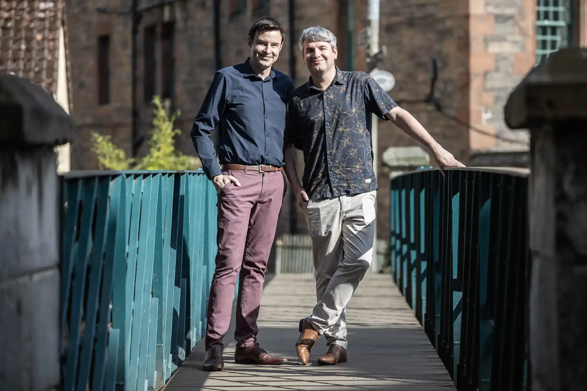 Two men stand side by side on a wooden bridge with teal railings, smiling at the camera. Brick buildings are visible in the background.