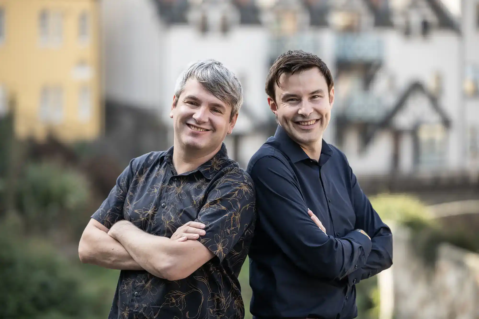 Two men stand outdoors, smiling with their arms crossed, in front of a blurred background of buildings.