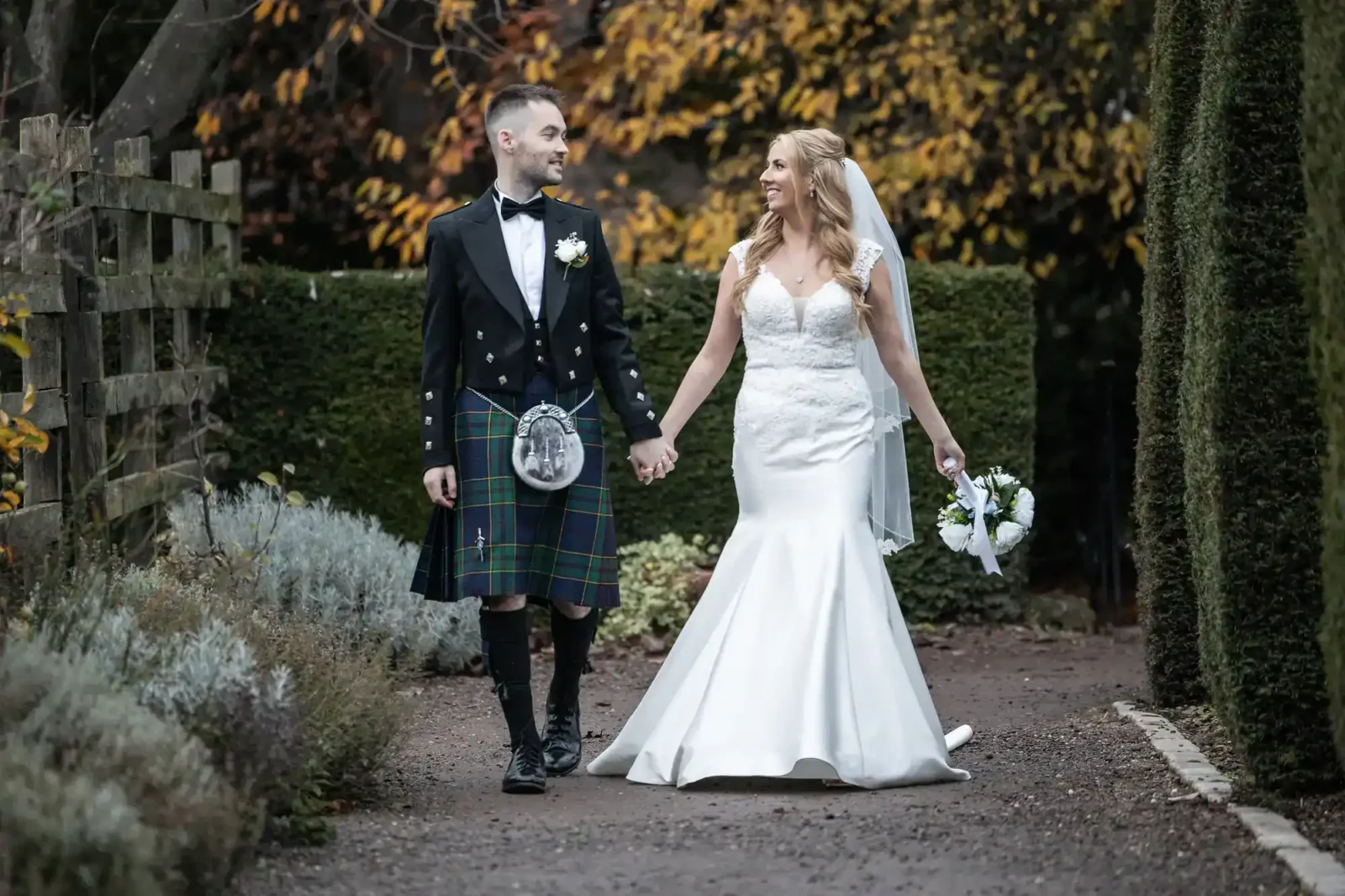 A bride in a white gown and a groom in a kilt hold hands and walk outdoors on a path, surrounded by greenery and autumn foliage.