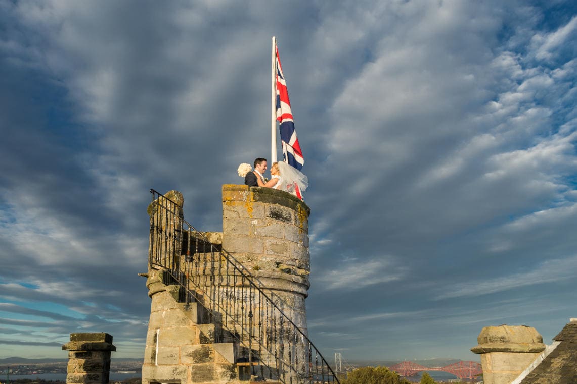 newlyweds embrace next to the Union Jack on the roof of the Auld Keep at Dundas Castle