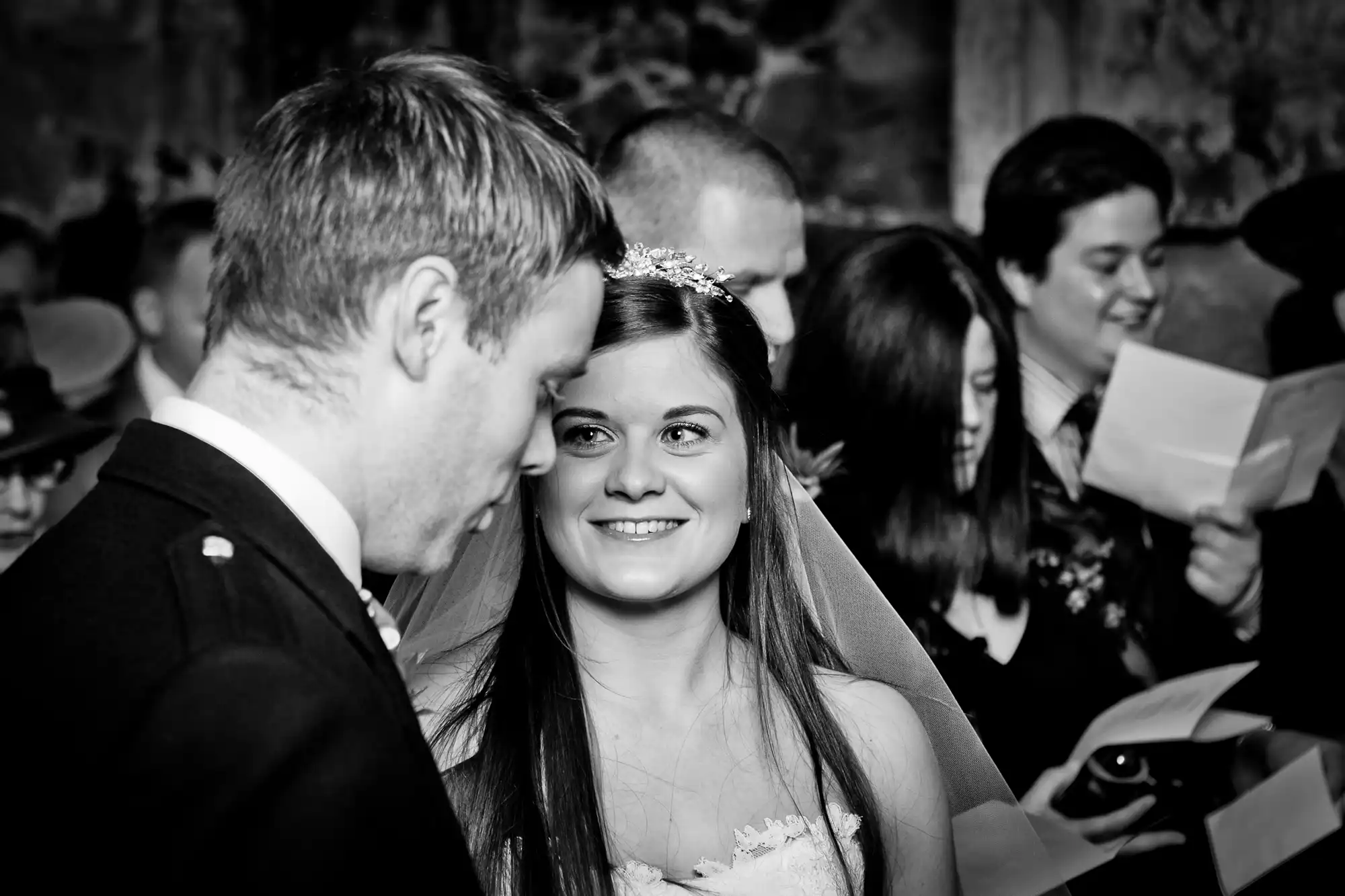 A bride smiling at her groom during their wedding ceremony, surrounded by guests in a dimly lit setting.
