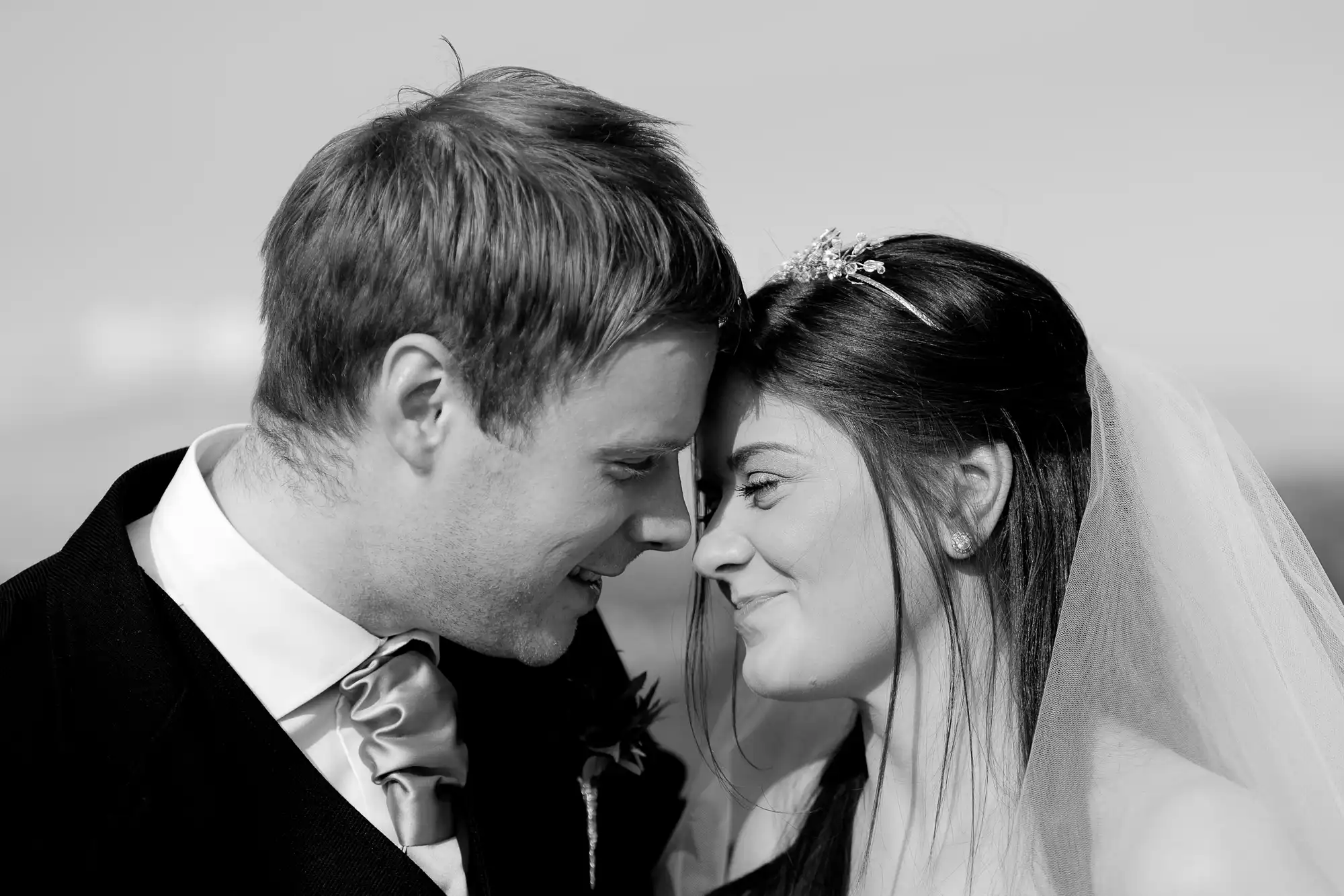 A black and white photo of a bride and groom smiling at each other, the bride wearing a veil and the groom in a tuxedo with a bow tie.