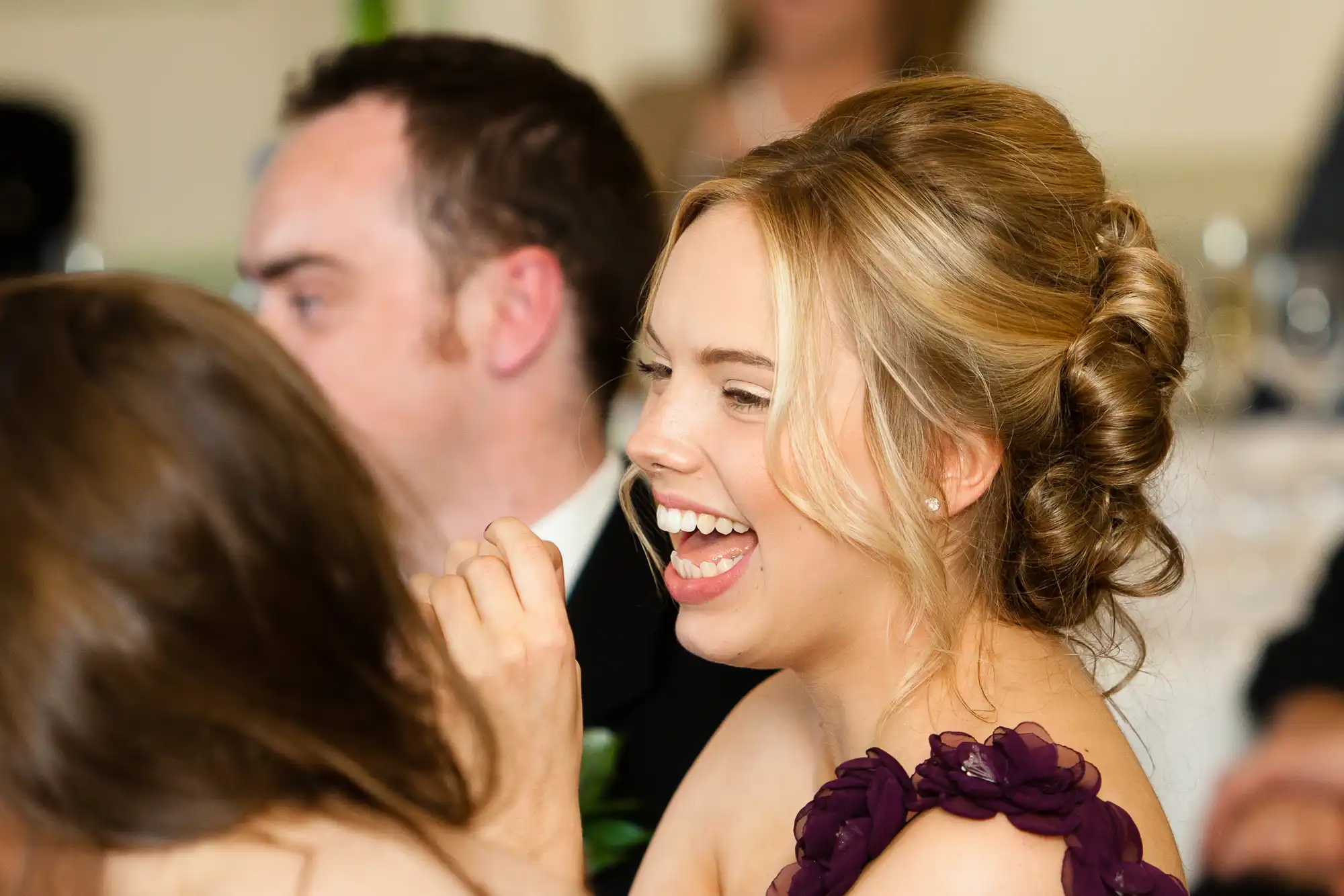 A woman laughing joyfully at a social event, dressed in a purple dress with floral details on the shoulder.