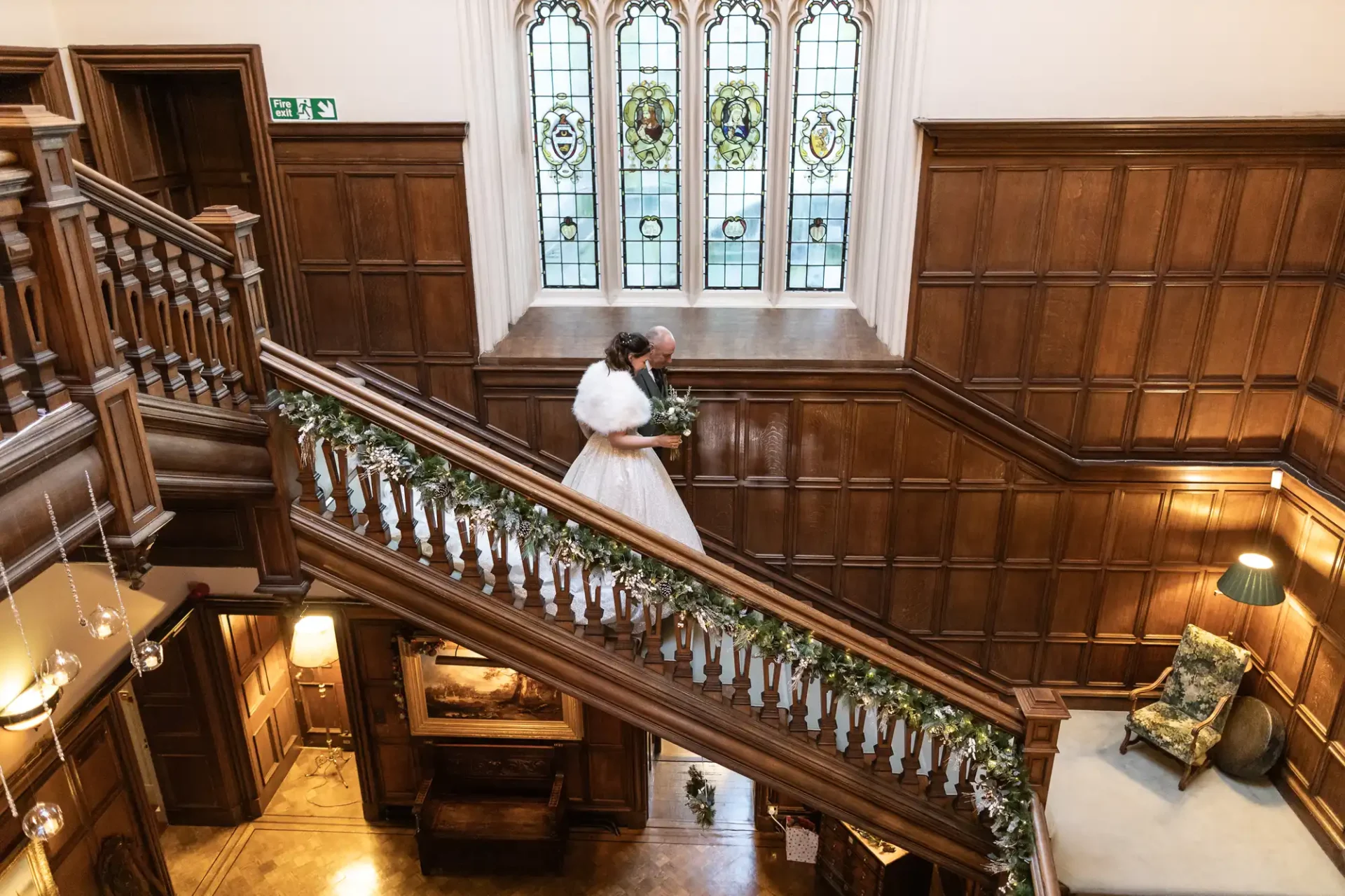 A bride and her father descend a wooden staircase adorned with greenery in a large, ornately decorated hall with stained glass windows.
