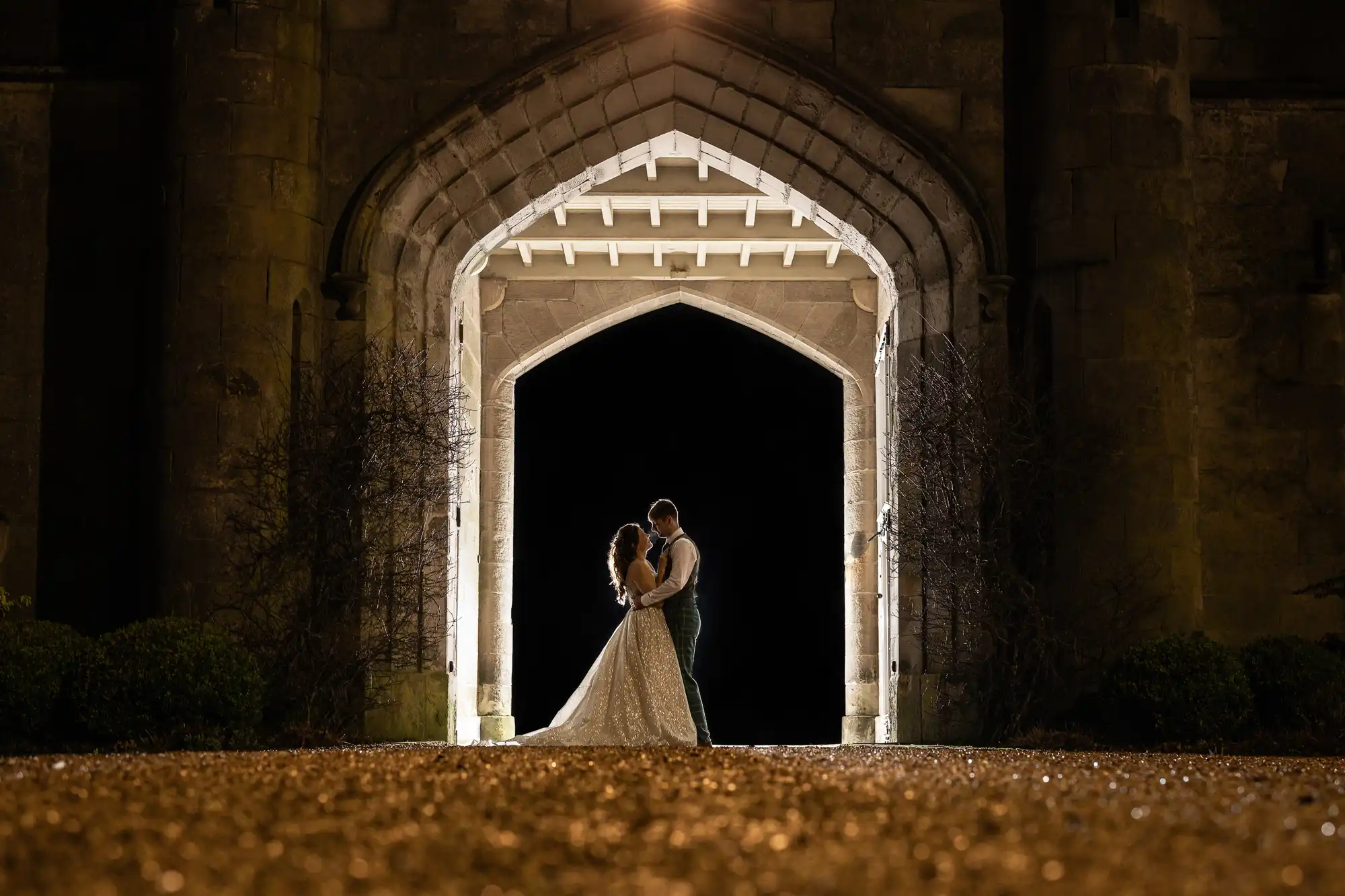 Dundas Castle wedding photography - capturing real moments at historic Scottish castles. A couple stands embraced under a gothic archway at night, illuminated by lights behind them, creating a silhouette against the dark background.