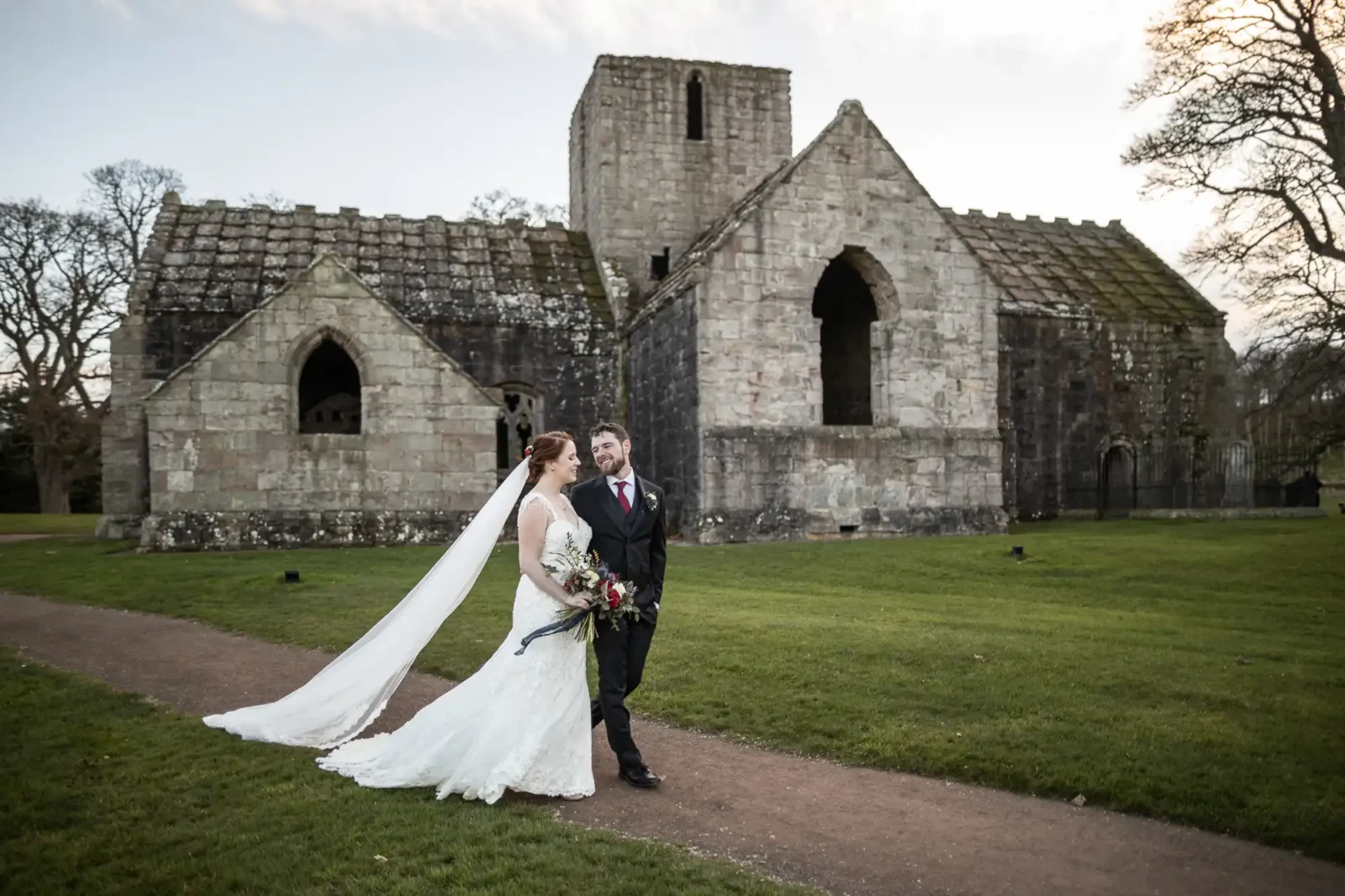 A bride and groom walk together outside an old stone church, Dunglass Church, with grass and trees in the background.