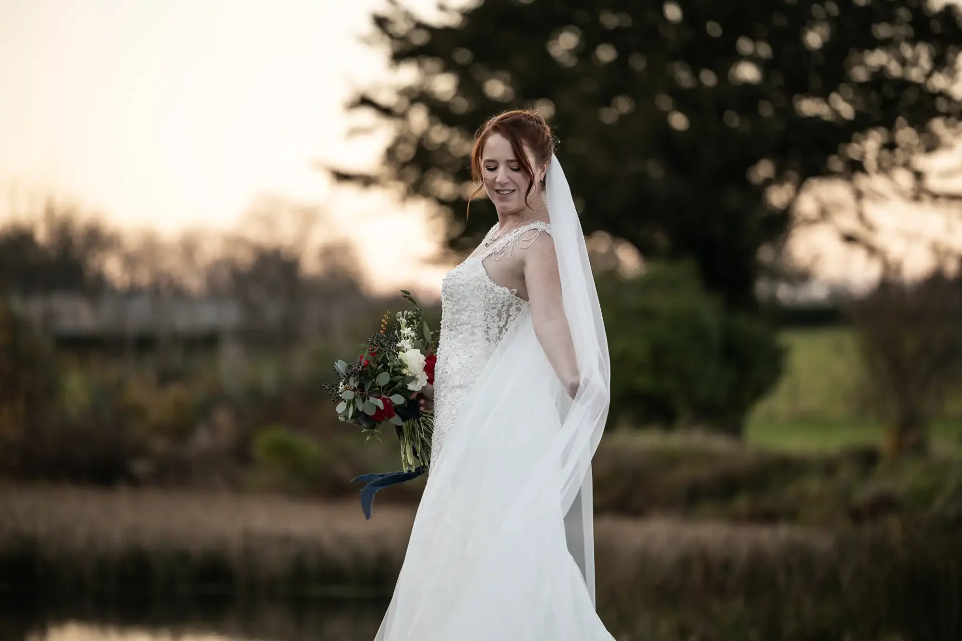 A bride in a white wedding dress and veil holds a bouquet outdoors, standing near water with greenery and trees in the background.