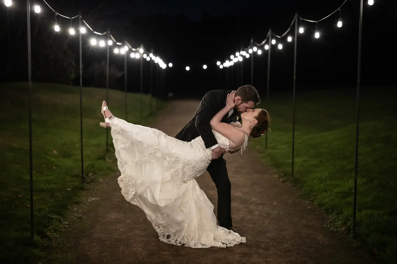Photography of a groom dips and kisses a bride in a white gown on a path lit by string lights at night, at Dunglass Estate, East Lothian.