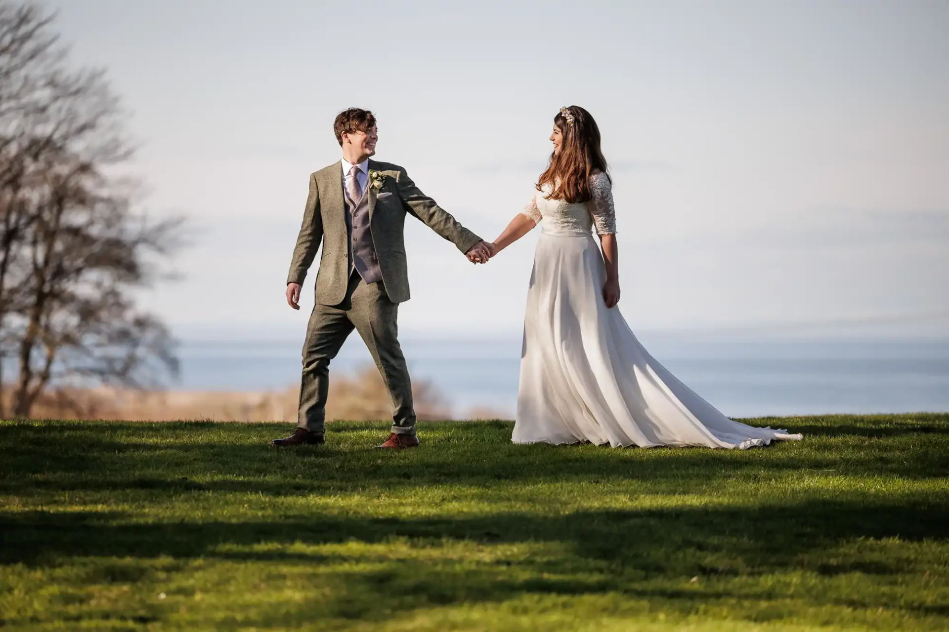 A bride and groom stand on grass, holding hands and looking at each other, with trees and water in the background at Dunglass Estate, East Lothian.