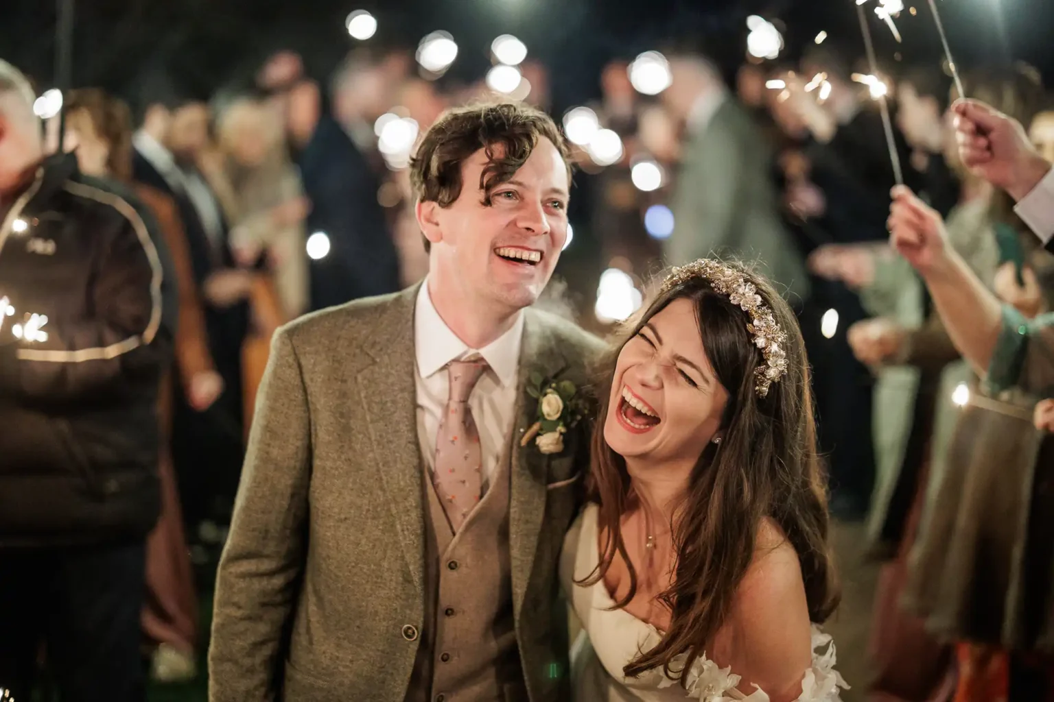 A newlywed couple in wedding attire laugh together while surrounded by guests holding sparklers at an outdoor celebration, beautifully captured by their wedding photographer at Dunglass Estate.