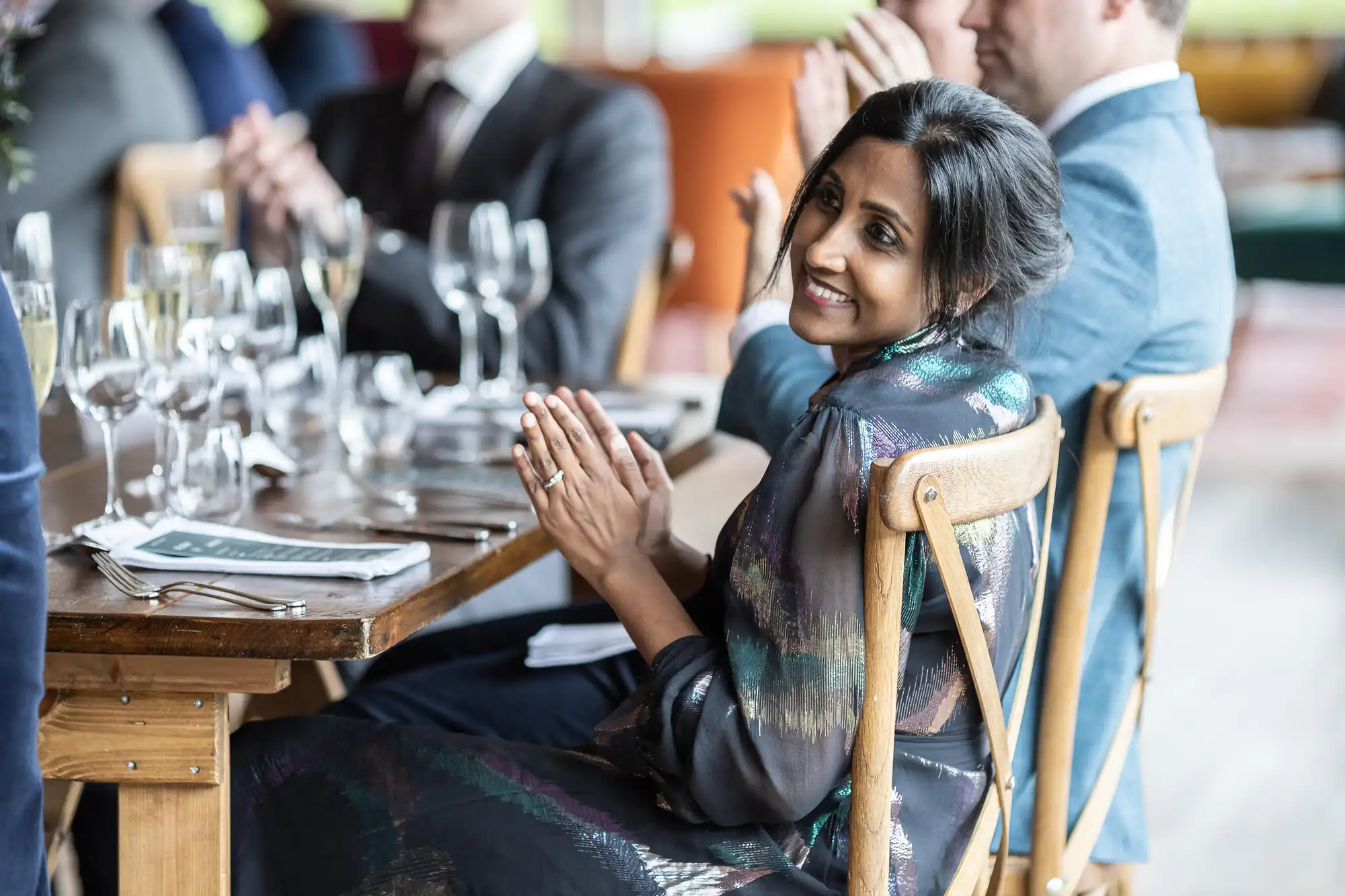 A woman in formal attire smiles and claps while seated at a table set with glassware and place settings, alongside other clapping individuals.
