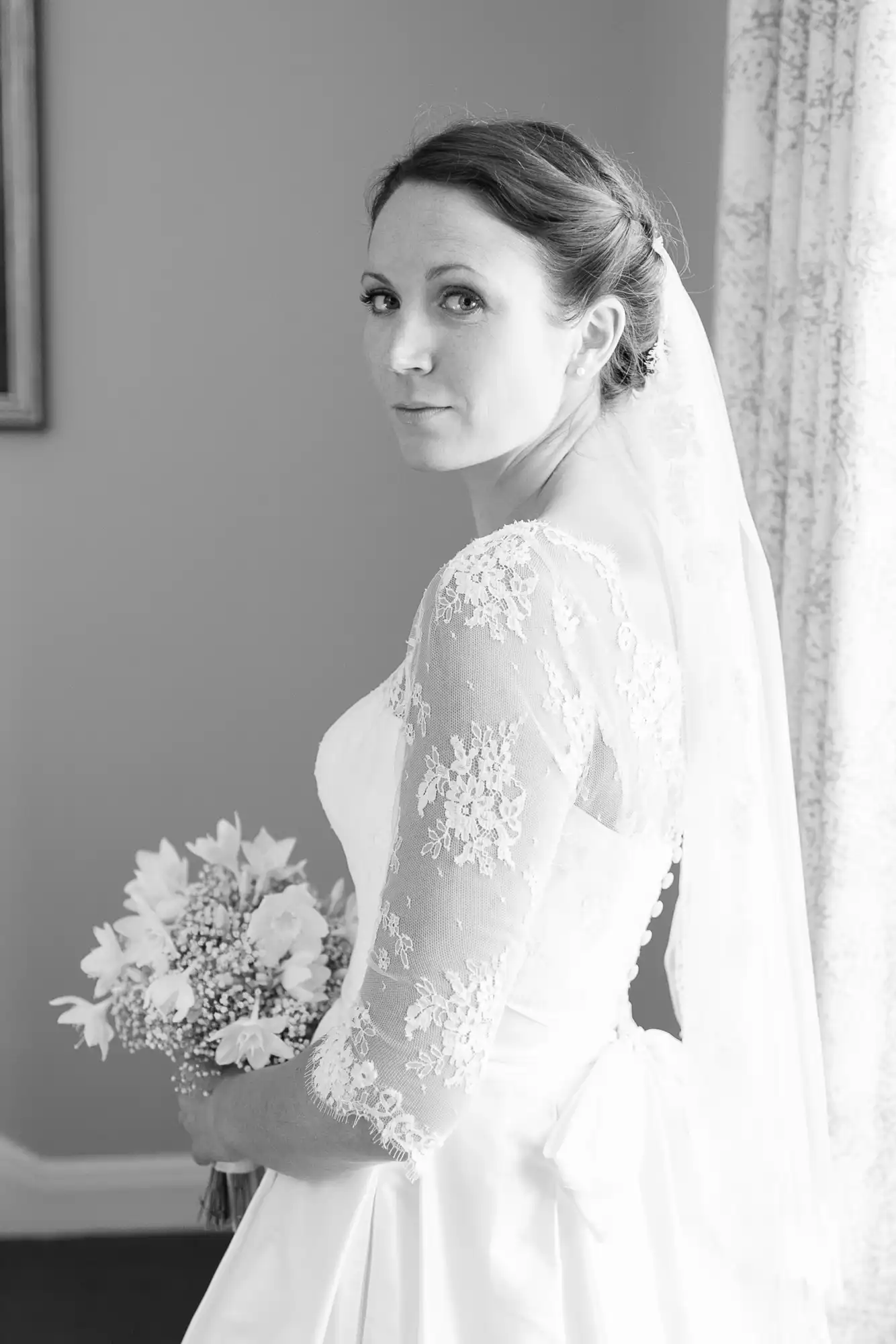 A bride in a lace wedding dress and veil holding a bouquet of flowers, looking over her shoulder in a black and white photo.