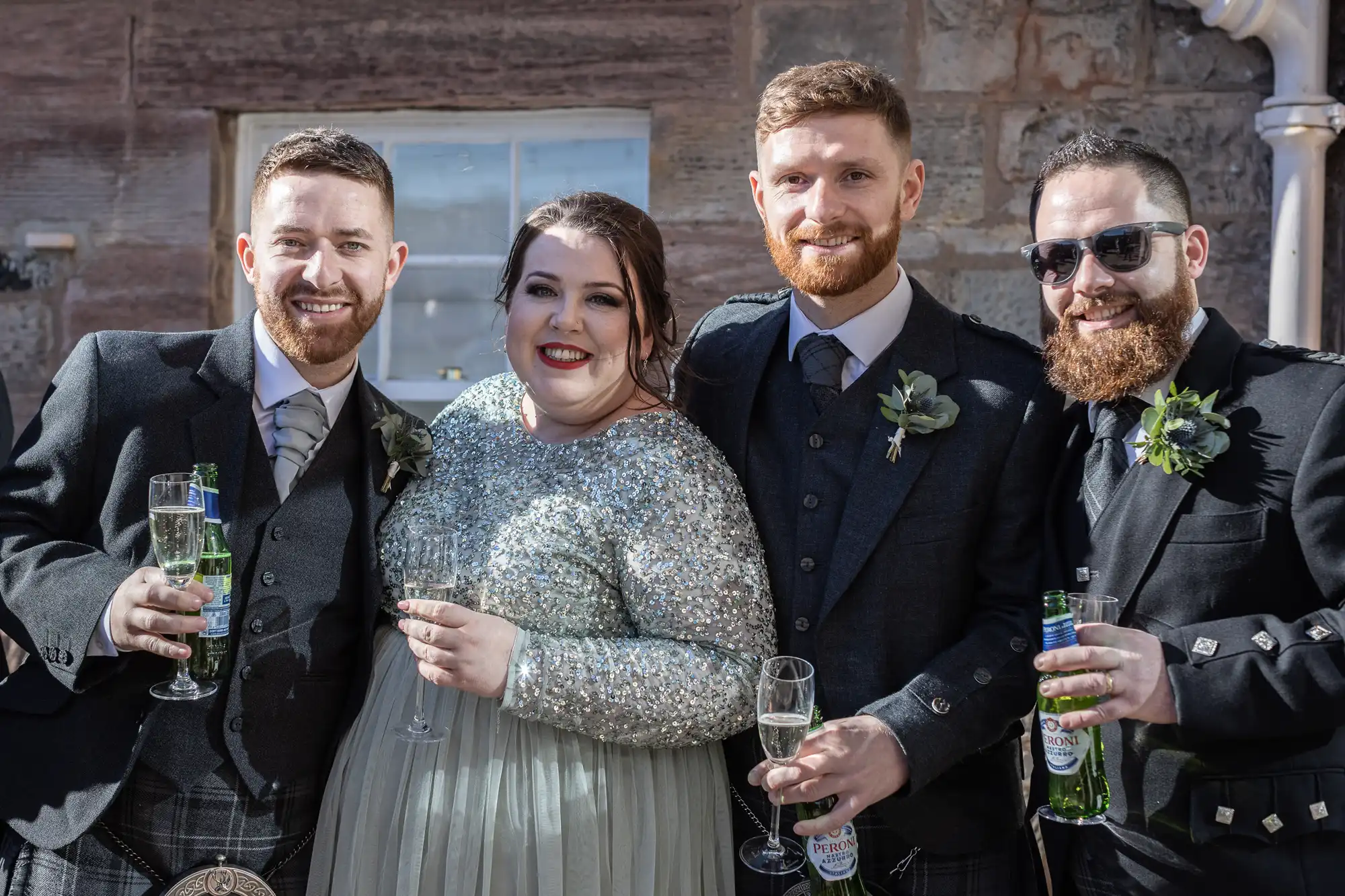 Four people dressed in formal attire standing together, three men and one woman, all holding beverage glasses. The group appears to be at a celebratory event, possibly a wedding, with a brick wall in the background.