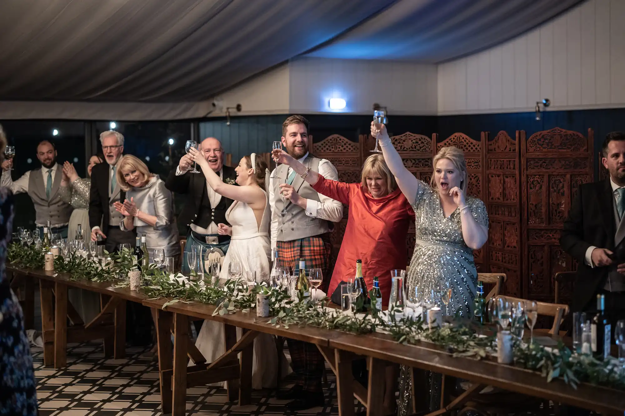 A group of people in formal attire stands and raises glasses in a toast at a wedding reception, with a long table decorated with greenery and candles in the foreground.