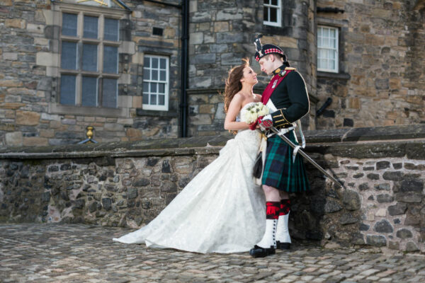 A bride in a white dress holding a bouquet smiles at a groom in a Scottish kilt and military uniform. They stand close together, leaning against a stone wall outside a historic stone building with large windows, during their Edinburgh Castle wedding.