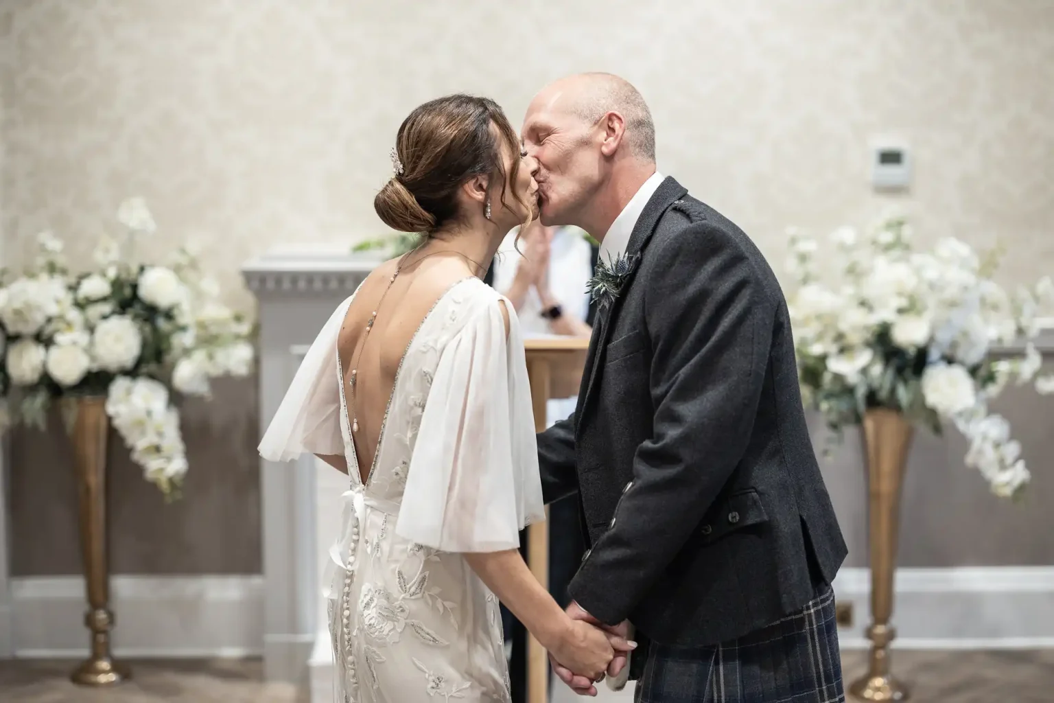 A bride and groom share a kiss at their wedding ceremony, holding hands. The bride is wearing a white dress, and the groom is in a dark suit. They are surrounded by elegant floral arrangements.