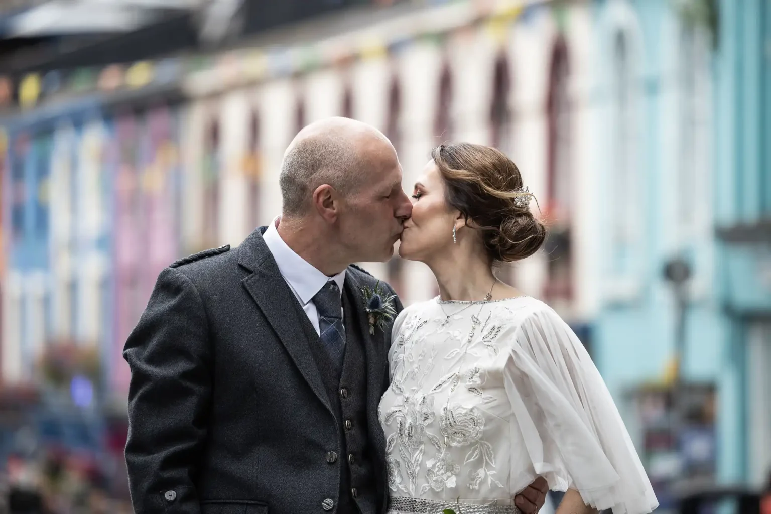 A couple kisses outdoors in front of a row of colorful buildings. The man is wearing a grey suit, and the woman is in a white dress with floral embroidery.