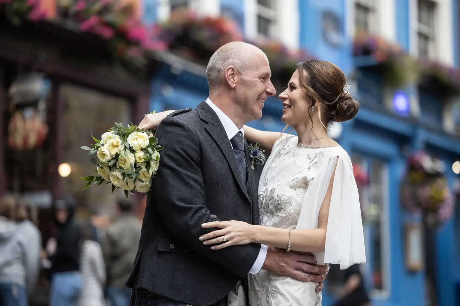 A couple in wedding attire shares a joyful moment outdoors. The bride holds a bouquet of white and yellow flowers. The background features a blue building adorned with hanging plants.