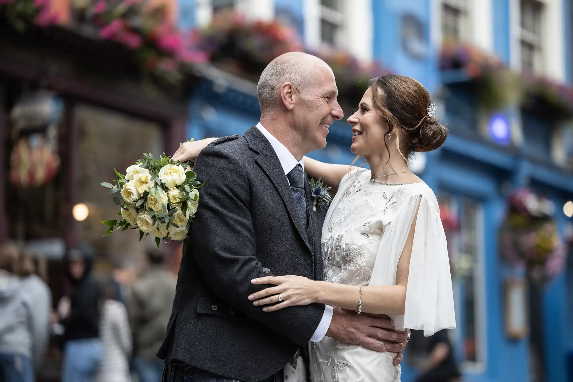A couple in wedding attire shares a joyful moment outdoors. The bride holds a bouquet of white and yellow flowers. The background features a blue building adorned with hanging plants.