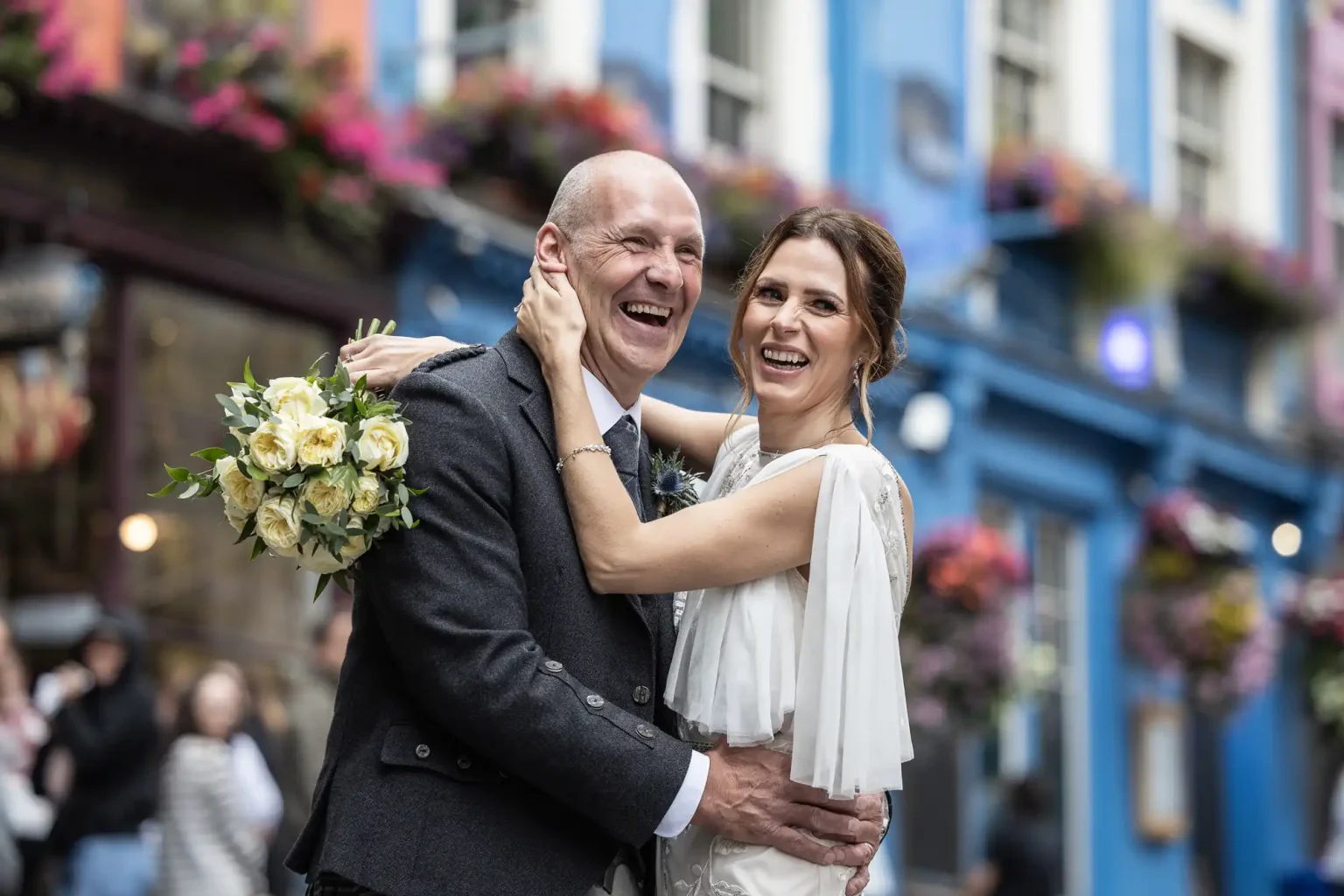 A smiling couple, dressed in wedding attire, stands close together in an outdoor setting with colorful buildings and flowers in the background. The man is holding a bouquet of white and yellow flowers during their City Chambers Old Town photos session