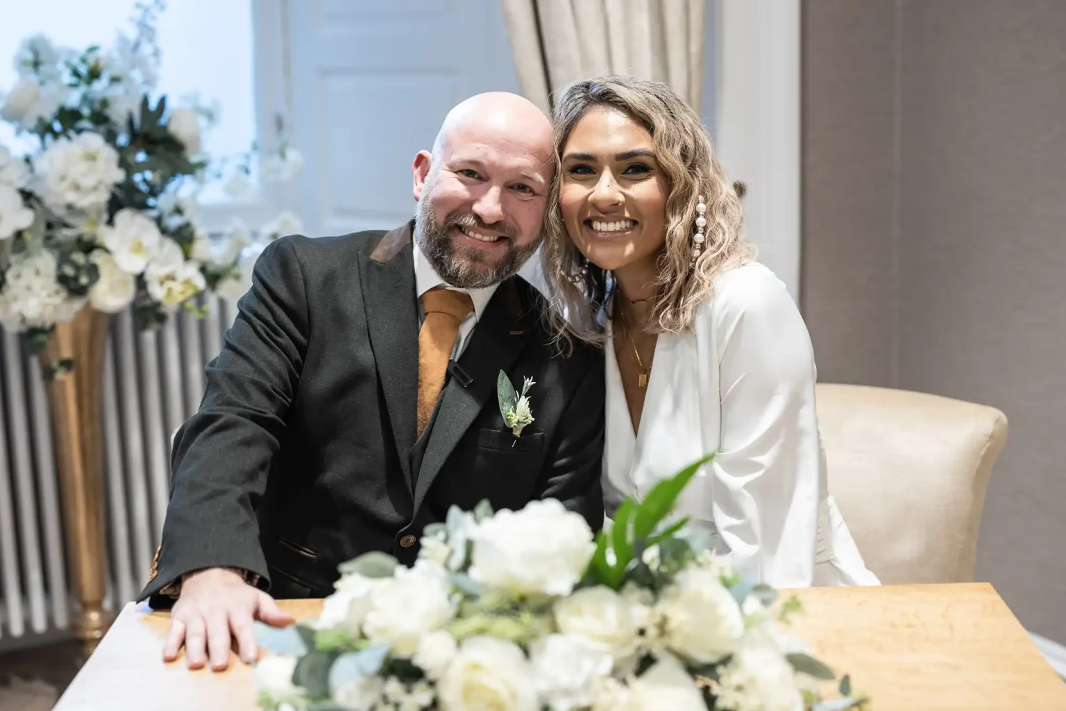 A couple poses together indoors at a table adorned with white flowers. The man wears a dark suit and tie, and the woman wears a white dress with her hair down. Both are smiling and sitting closely.