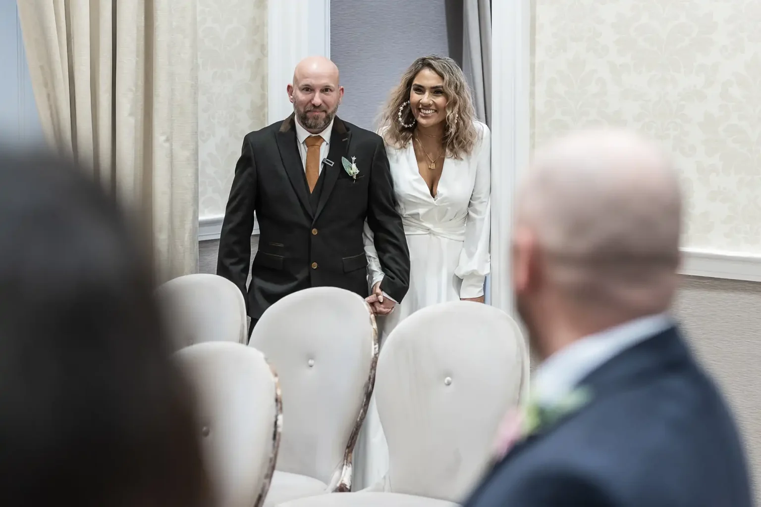 A couple holding hands enters a room with white cushioned chairs. The woman wears a white dress, and the man wears a dark suit, as they approach people facing them from the foreground.