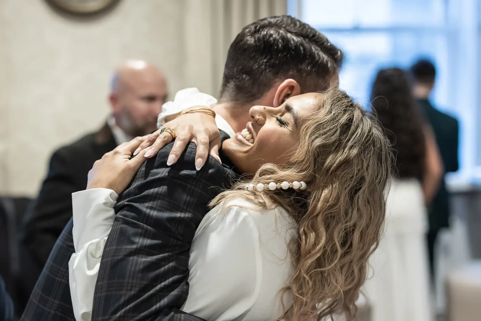 Two people share a joyful hug indoors. The woman has blonde hair and wears a white outfit with pearl earrings. Other people and a window are visible in the background.
