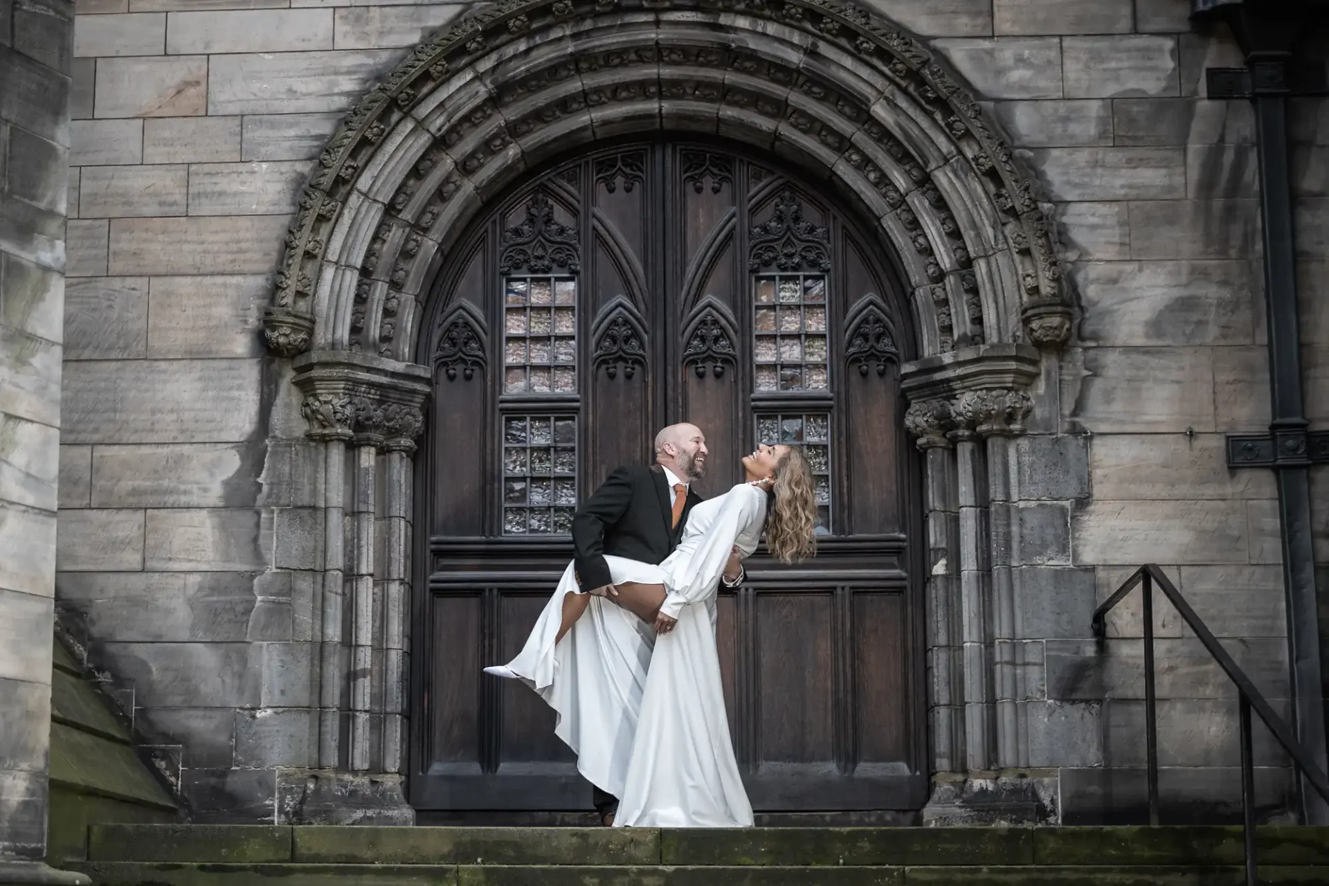 Edinburgh City Chambers weddings - the beautiful elopement of Sara and Carl - A man in a black suit and a woman in a white gown pose in front of an arched wooden door, with the man holding the woman in a playful dip.