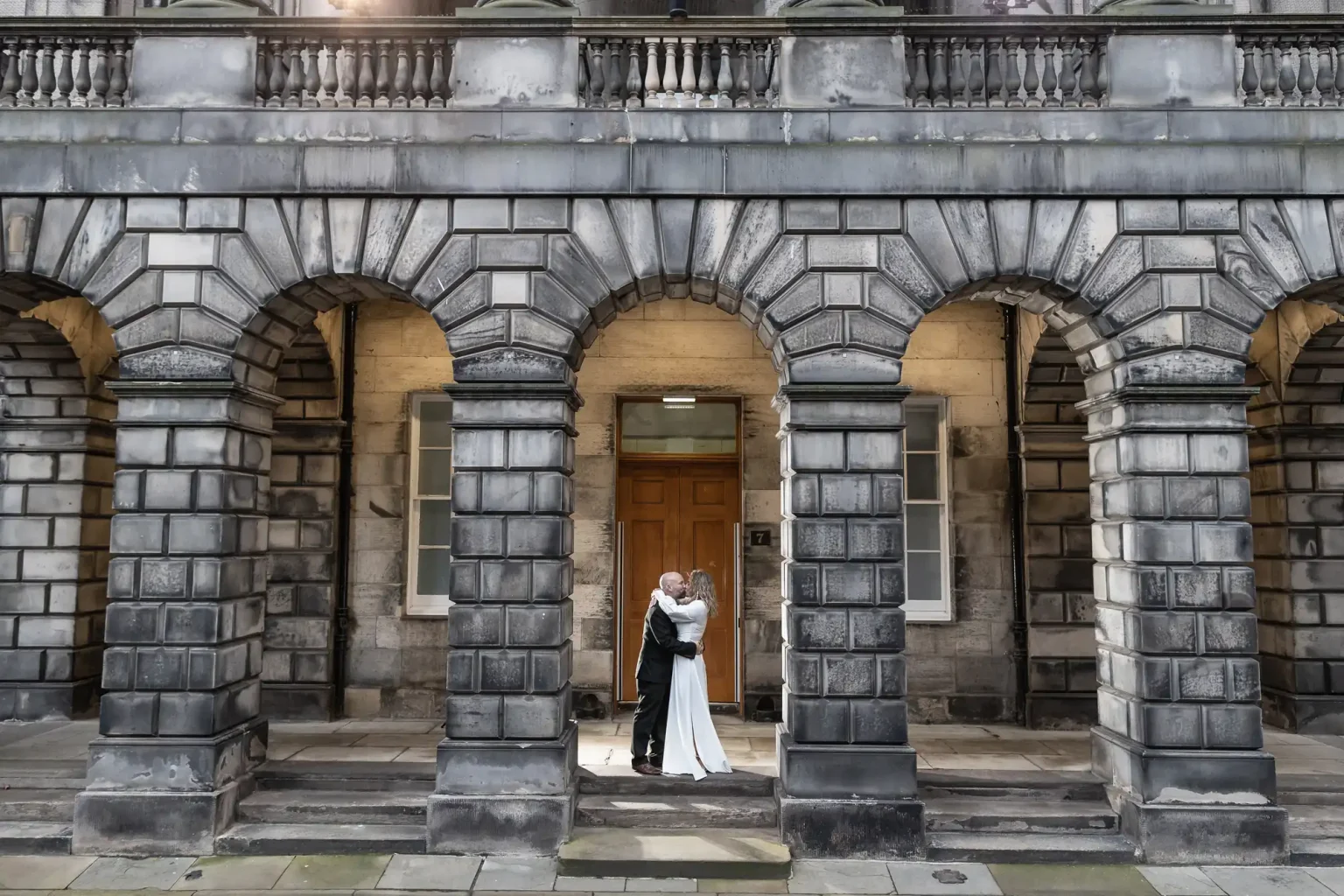 A couple dressed in wedding attire embraces under the archways of a historic stone building.