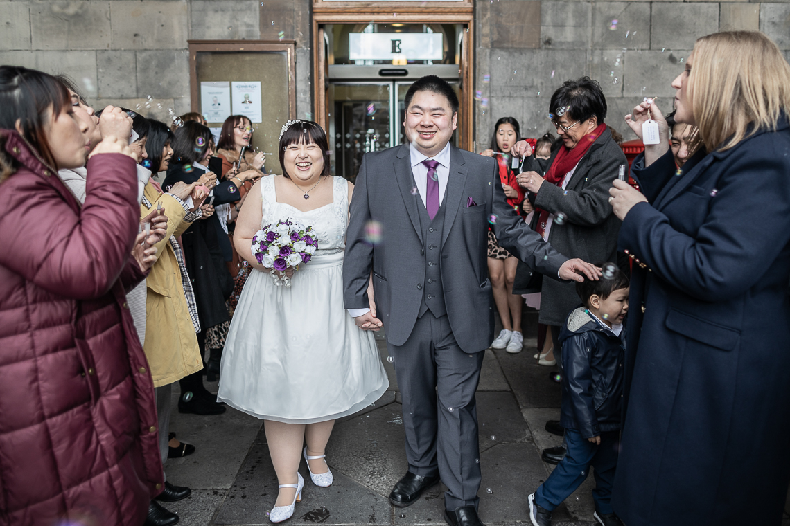 A bride and groom walk hand in hand out of a building surrounded by guests blowing bubbles and celebrating.