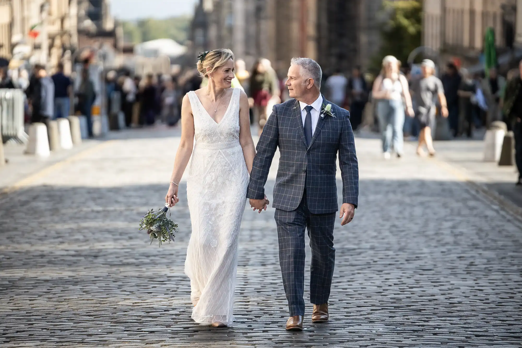 A bride and groom hold hands and walk down a cobblestone street, smiling at each other, with people in the background.