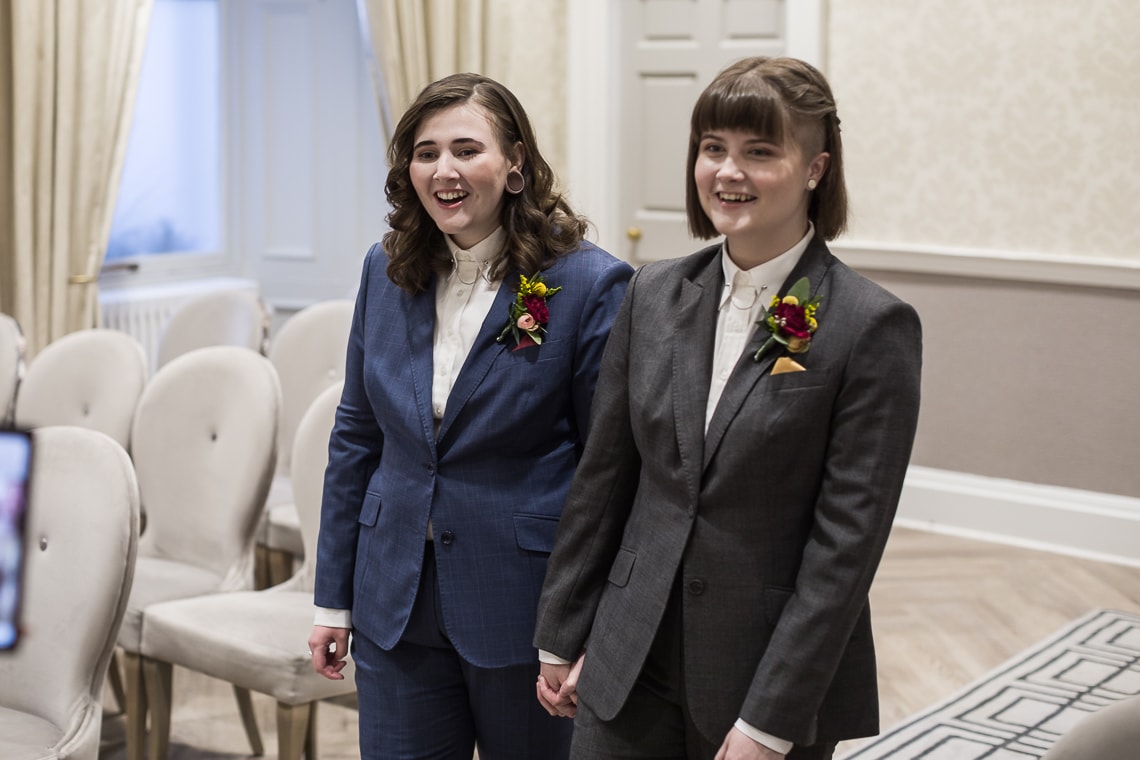 Two women in suits holding hands and smiling, standing in a room with empty chairs and neutral decor, possibly at a wedding or formal event.