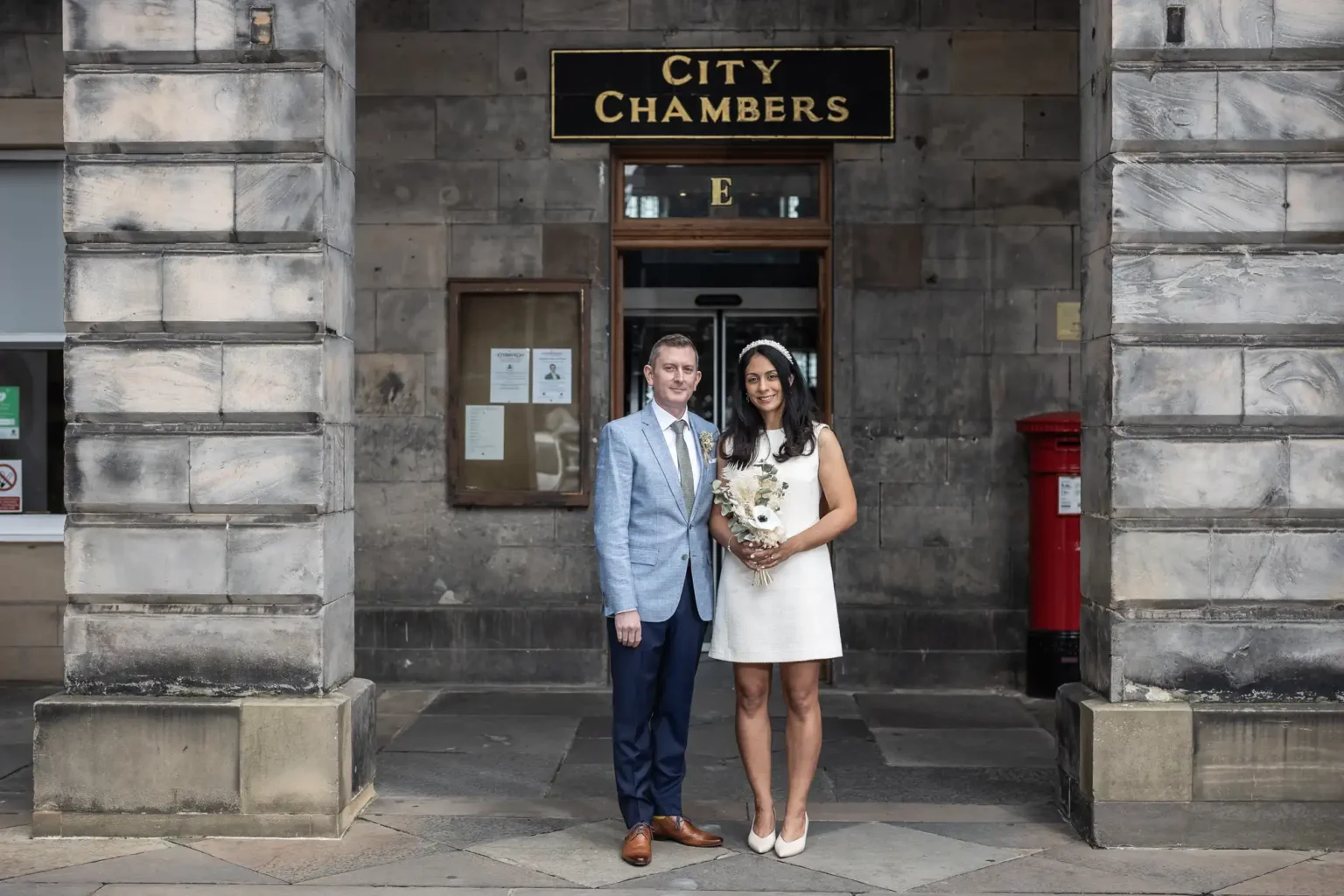 City Chambers summer elopement - a couple, dressed formally, stands in front of the City Chambers building entrance, with the woman holding a bouquet of flowers.