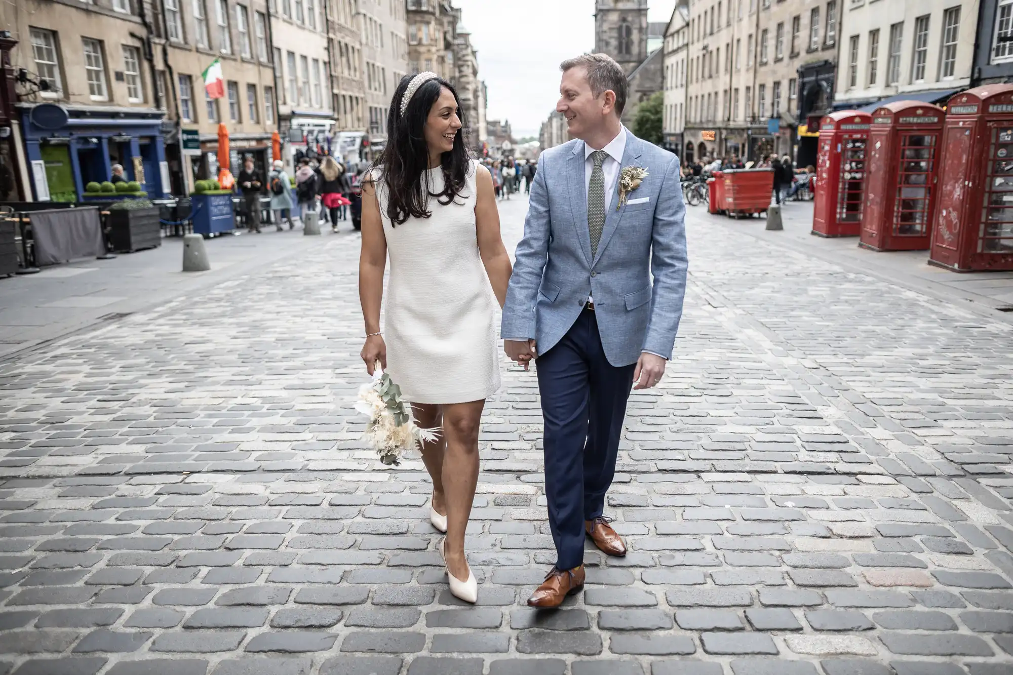 A couple in formal attire holds hands and walks down a cobblestone street. The woman wears a white dress and carries a bouquet. The man wears a blue blazer and tie. Red phone booths and buildings line the street.