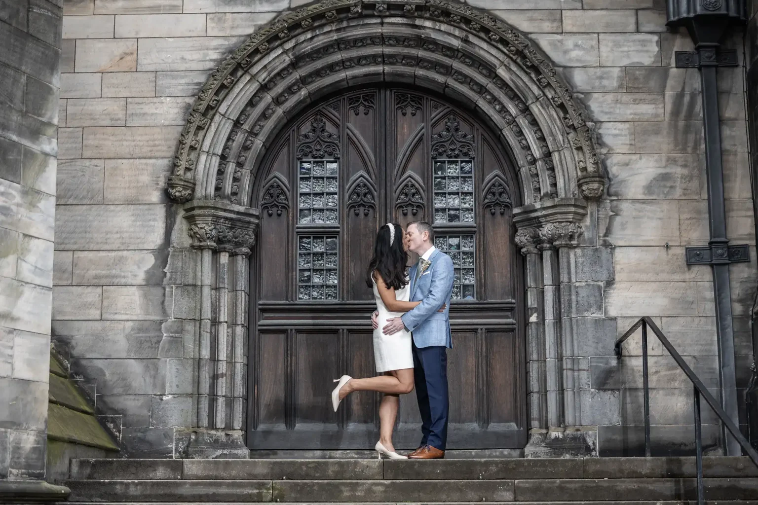 A couple shares a kiss in front of an ornate, arched wooden door of a stone building. The woman is lifting one leg and wearing a white dress, while the man is in a light blue blazer and dark pants.