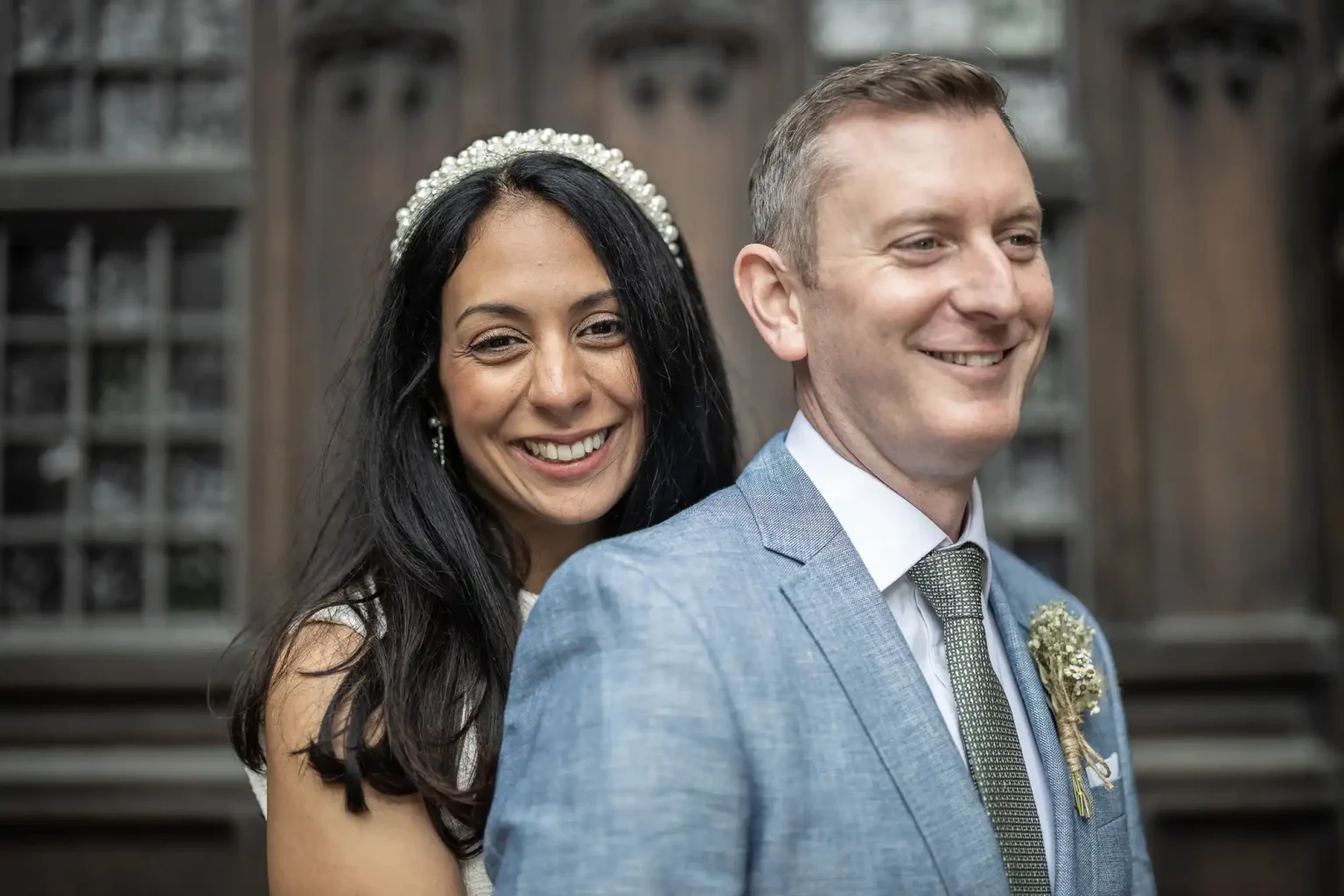 A woman with long dark hair and a headband stands smiling behind a man in a light blue suit with a boutonniere, outside a historic building.