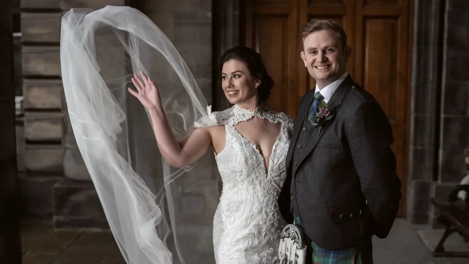 A bride in a white gown waves as her veil flows in the air, standing next to a smiling groom in a suit with a tartan kilt, in front of wooden doors.