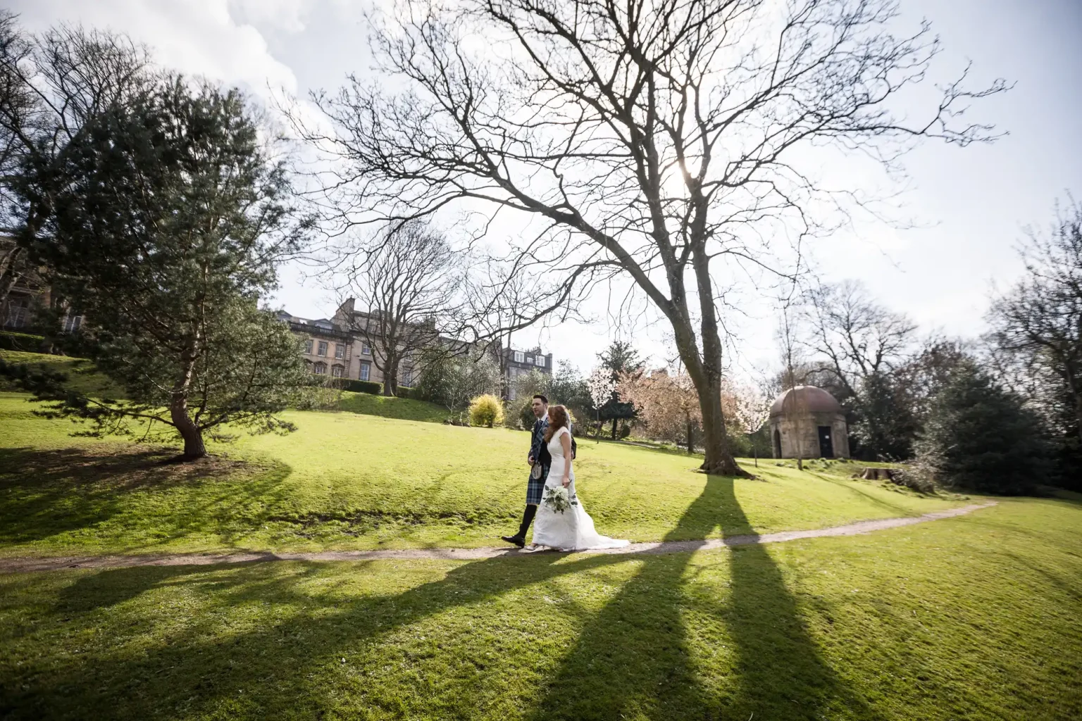 Colour wedding photography of a bride and groom walking hand in hand on a narrow path through a sunlit Queen Street Gardens park with trees and grass, shadows stretching across the ground, and buildings in the background.