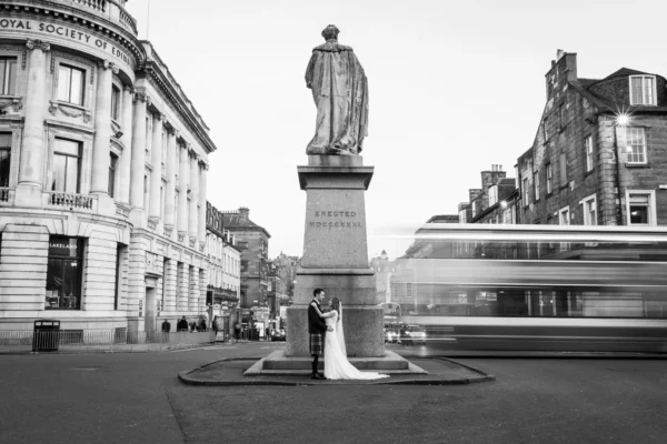 Karyn and Christopher’s InterContinental Edinburgh The George wedding. A bride and groom stand together at the base of a large statue in a city junction. Motion blur from a passing bus appears on the right. Surrounding Victorian buildings and a nearly empty street are visible. The photo is in black and white.