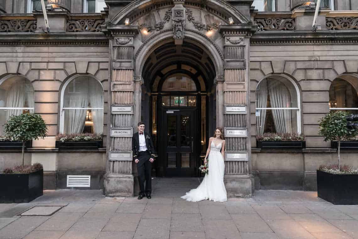 George Hotel newlyweds Paula and Alex: A couple, dressed in formal wedding attire, stands in front of an ornate building entrance. The groom is on the left, leaning against a pillar, and the bride is on the right, holding a bouquet.