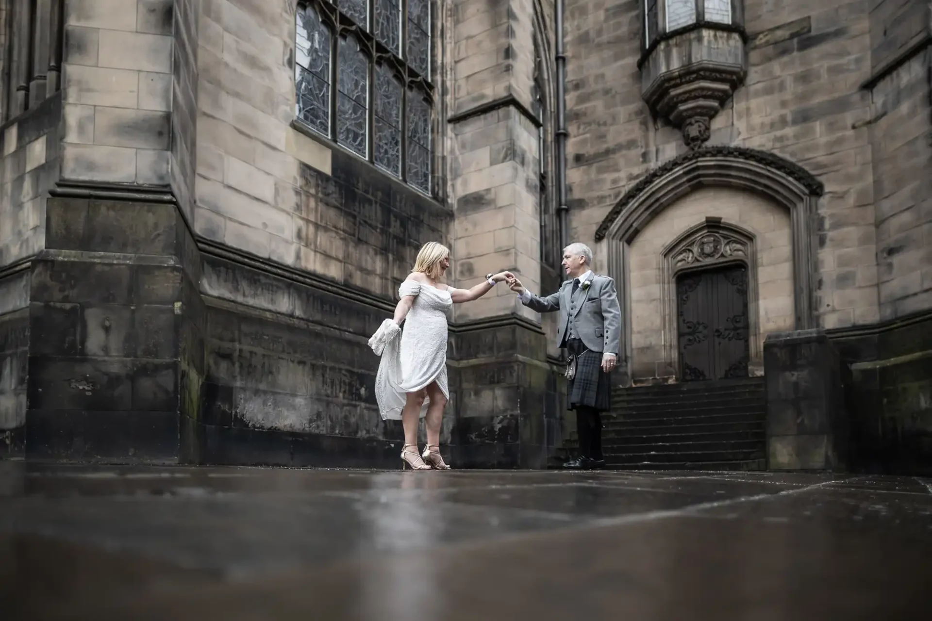 A couple dressed in formal attire holds hands and poses outside a large stone building with arched windows and doors.