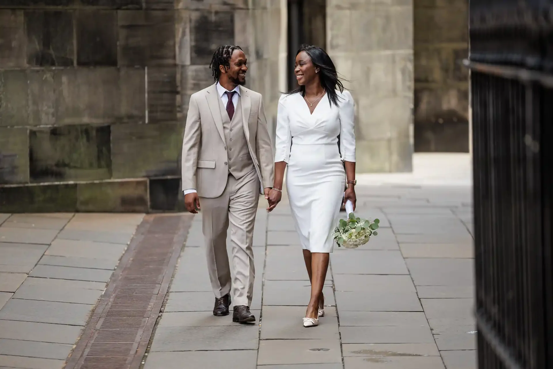 A couple dressed in formal attire, holding hands and walking outdoors on a stone pathway at Parliament Square during their Edinburgh elopement. The woman holds a bouquet of flowers.