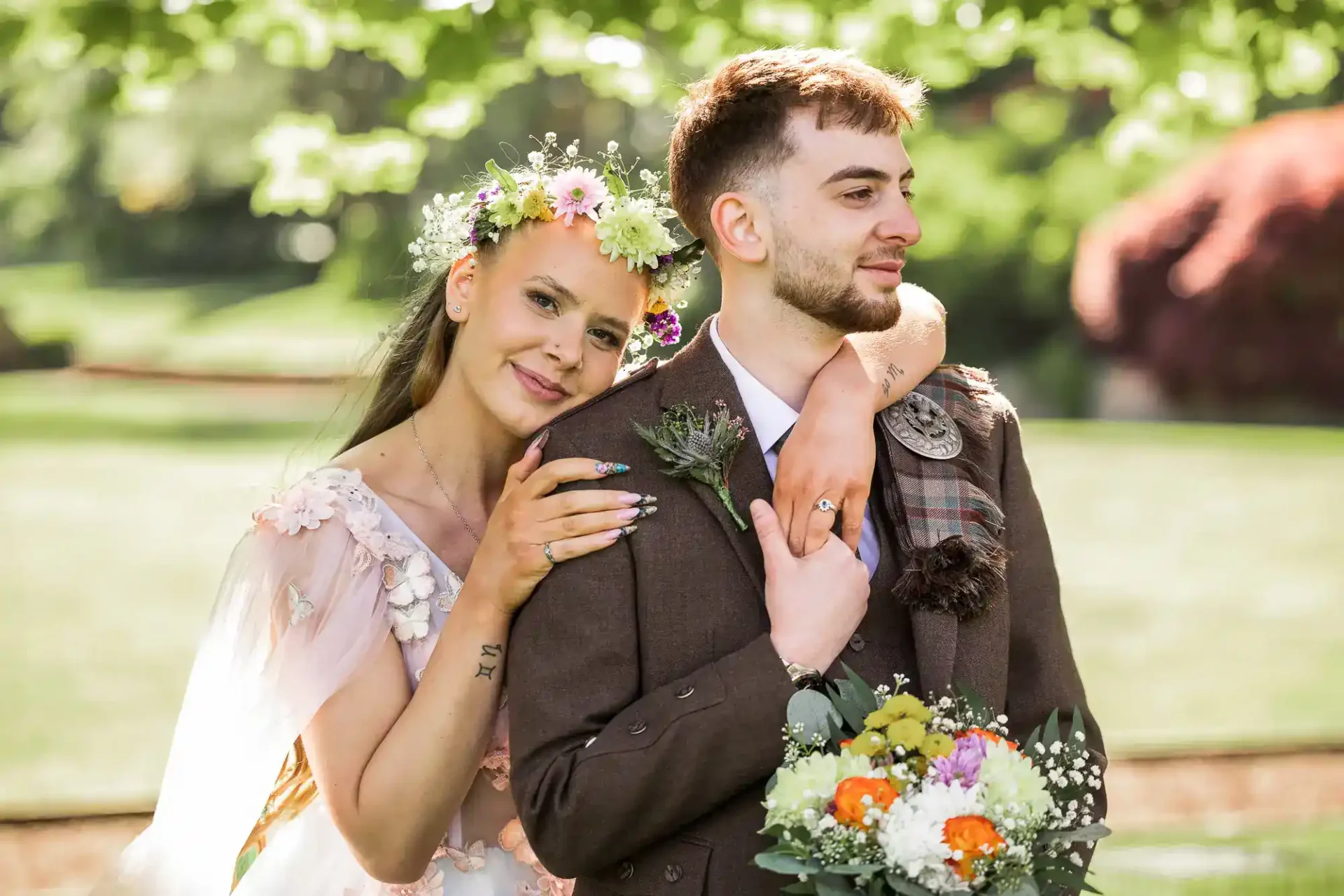 Edinburgh wedding advice hub,. A bride with a flower crown and a groom in a brown suit stand together outdoors, with the bride resting her chin on the groom’s shoulder and holding a bouquet of flowers.