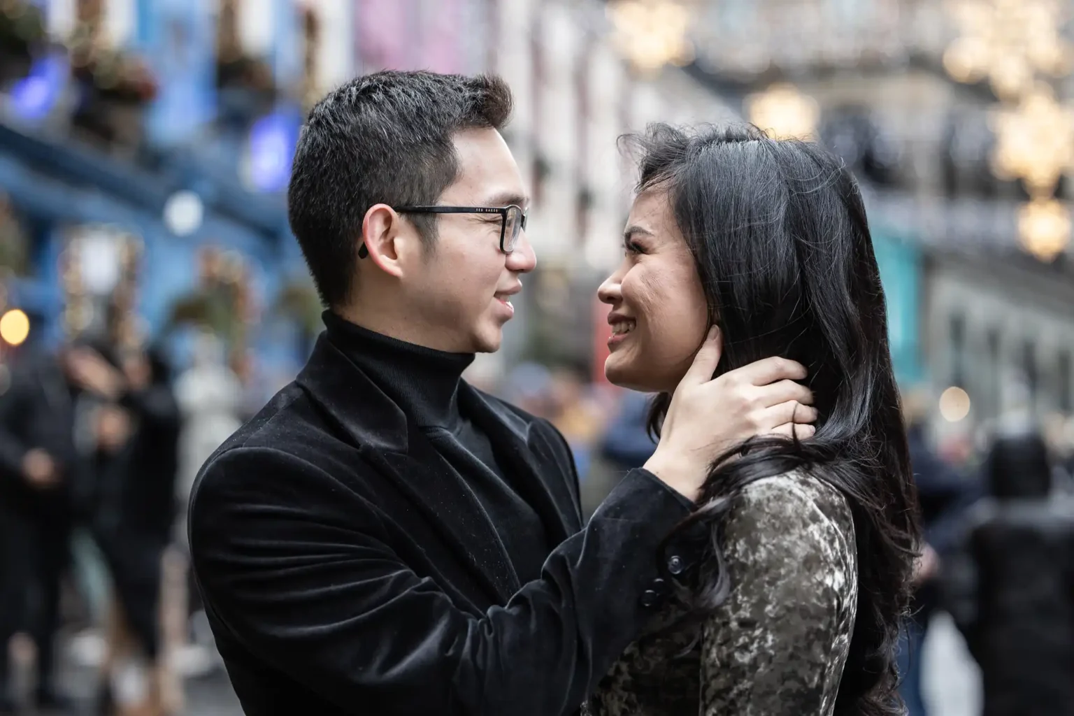 A man and woman smiling at each other while the man gently holds the woman's neck. They are outdoors with blurred buildings and lights in the background.