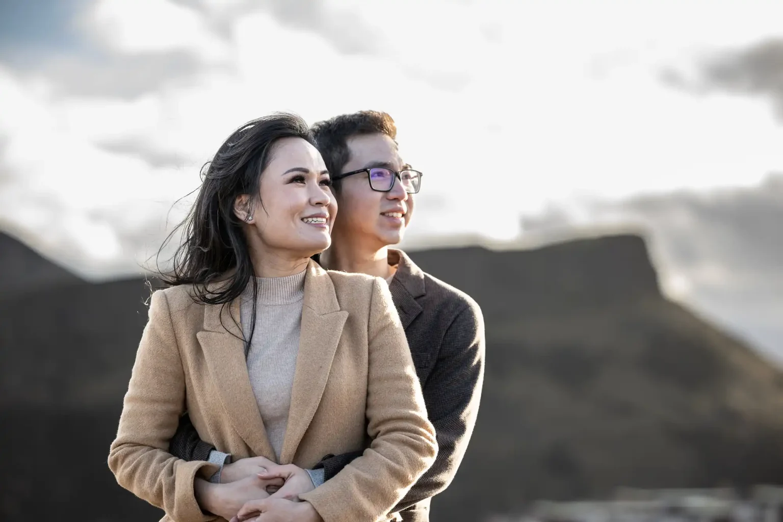 A couple standing outdoors, with one person hugging the other from behind. Both are looking off-camera. The background features a blurred mountainous landscape under a cloudy sky.