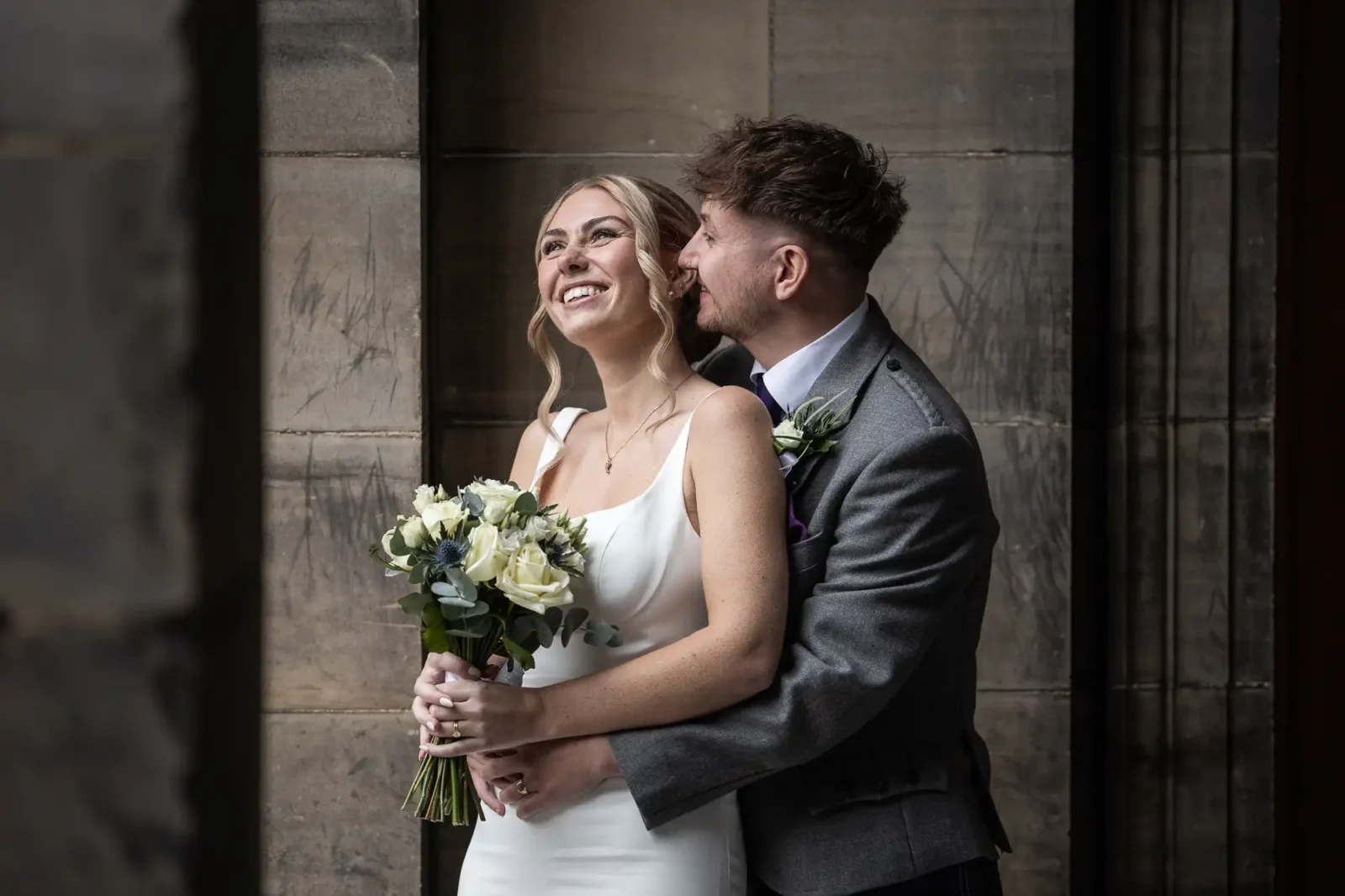A bride holding a bouquet smiles while the groom stands behind her, embracing her and kissing her cheek in front of a stone wall.