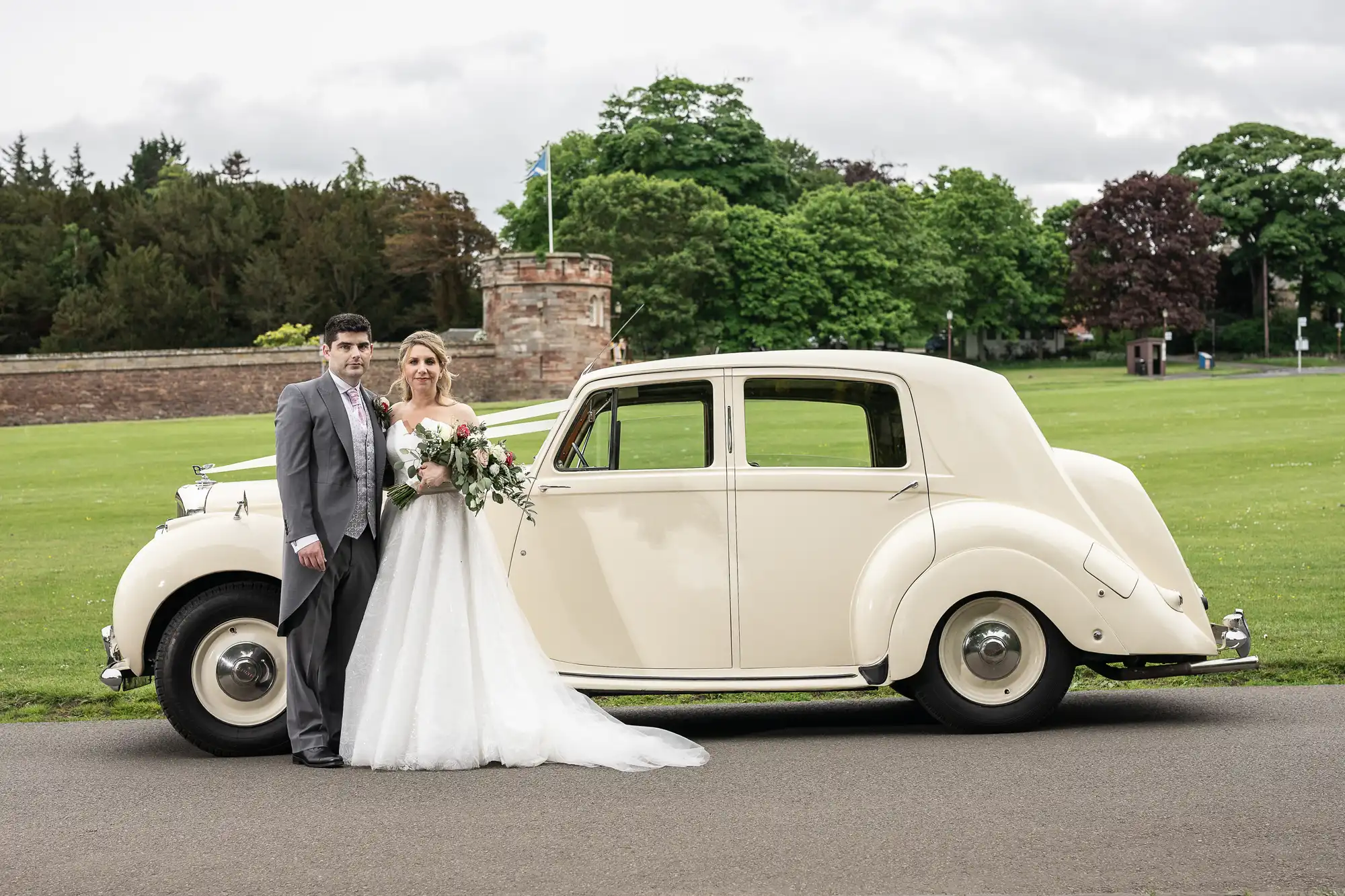 Dirleton Kirk wedding photographer: A bride and groom stand next to a vintage cream-colored car, posing for a photo in front of a grassy area with trees and a stone structure in the background.