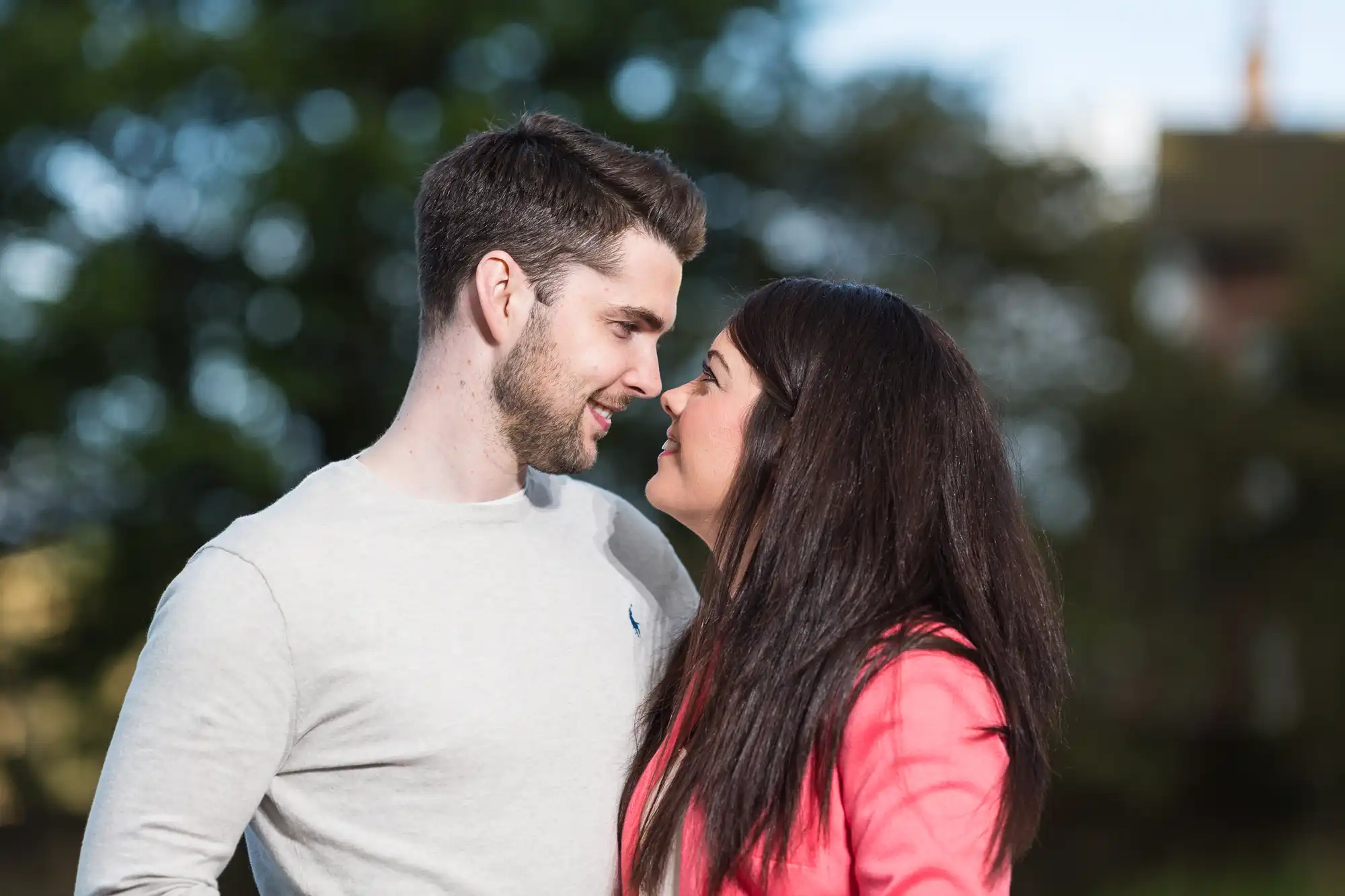 A young couple smiling at each other lovingly, with blurred greenery in the background. the man is in a gray shirt, and the woman wears a bright red jacket.
