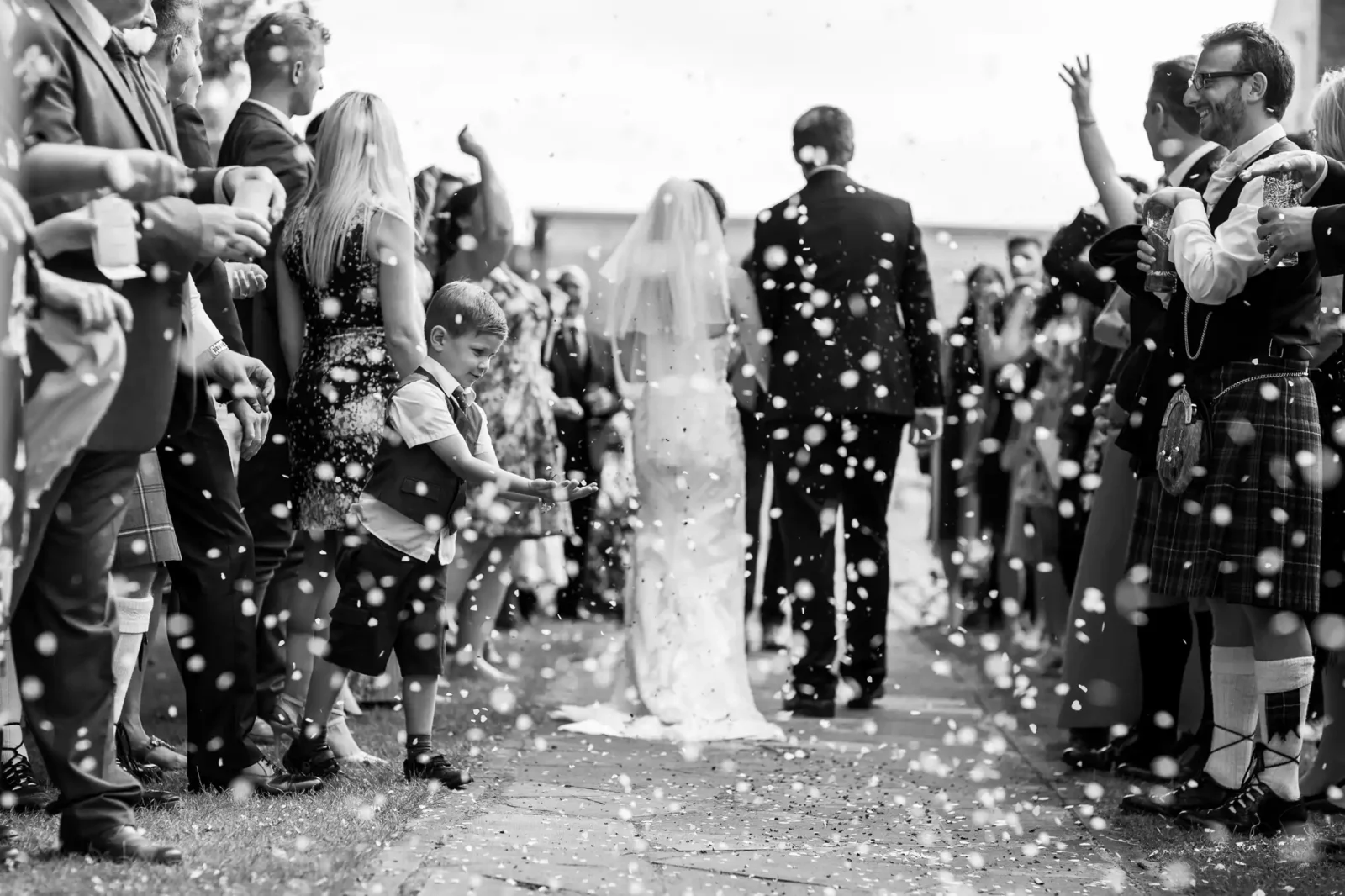 Black and white wedding photo image of a wedding celebration with guests throwing confetti as a bride and groom walk down an outdoor aisle at The Venue Eskmills, Musselburgh, East Lothian.